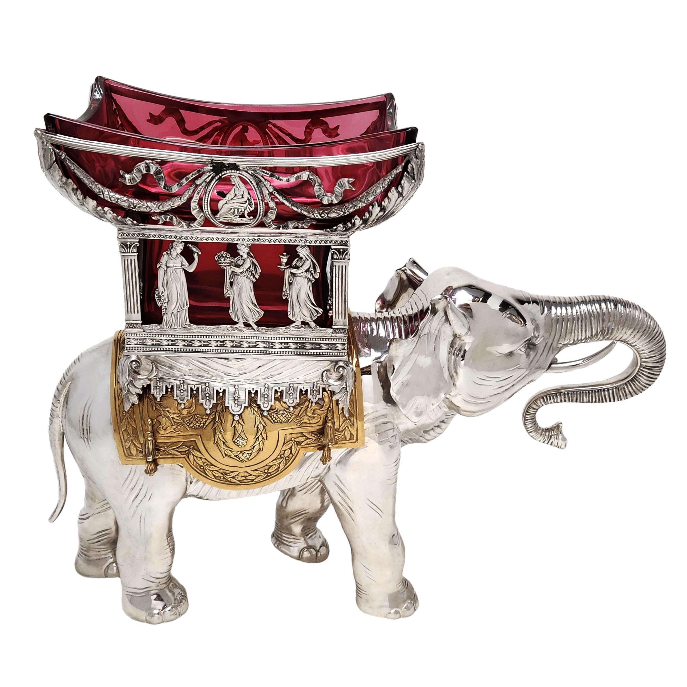 A magnificent solid silver elephant centrepiece supporting a gilt saddle with a large red glass bowl in a pierced Silver Basket. The basket features a band of classical figures and a swag and bow design with acanthus leaf corners and chased floral