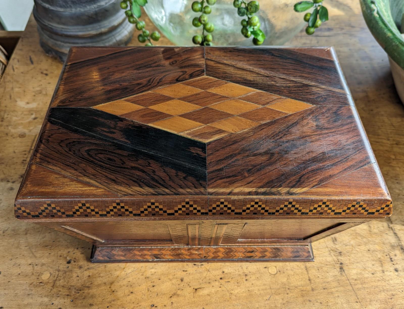 A finely crafted antique wood marquetry box with hinged lid and green fabric interior. This box has a refined geometric design featuring both checkered and diamond shaped patterns on a sarcophagus shape. Measures 12.5 inches in width by 7.5 inches