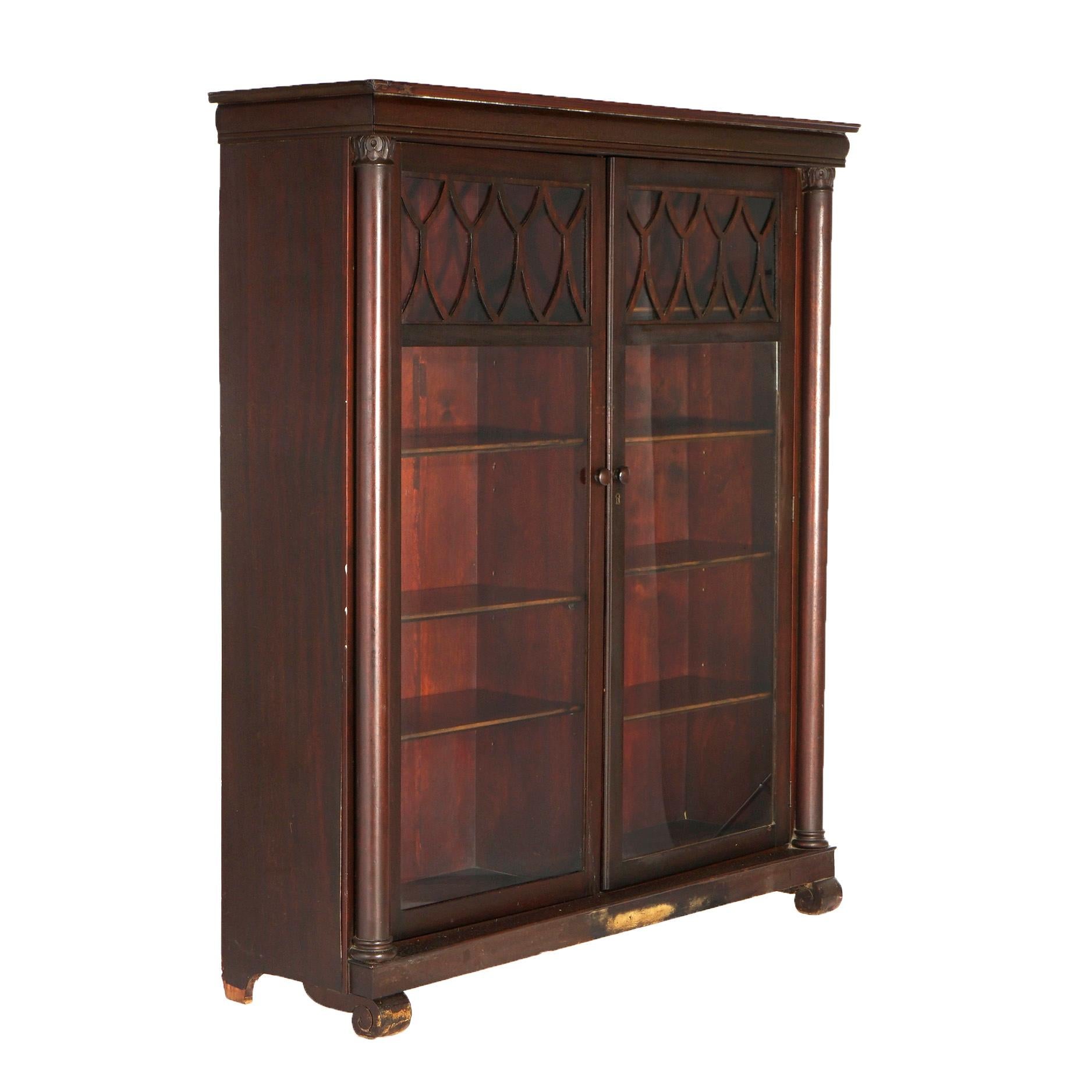 An antique Classical Greco American Empire bookcase by Larkin offers mahogany construction with double glass doors having lattice facing and opening to a divided adjustable shelf interior, flanked by full columns having carved acanthus capitals,