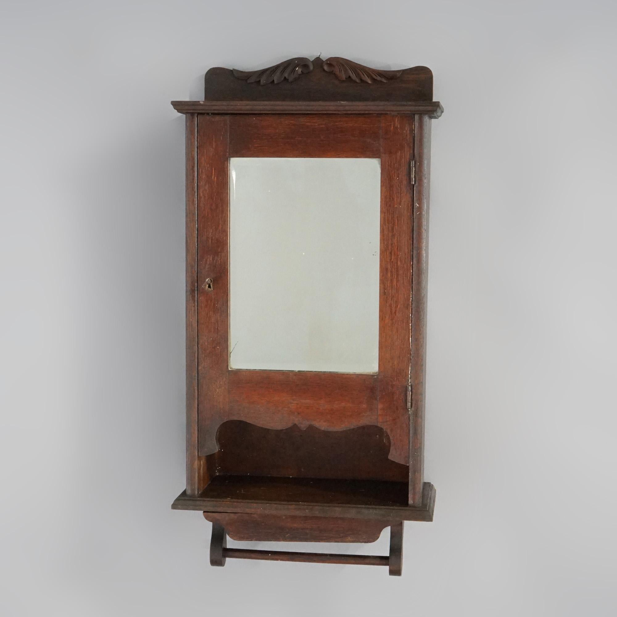 An antique Larkin medicine wall cabinet offers oak construction with shaped backsplash having carved foliate elements over case with single mirrored door opening to compartmentalized interior, over towel bar and small pull-out drawer, working lock