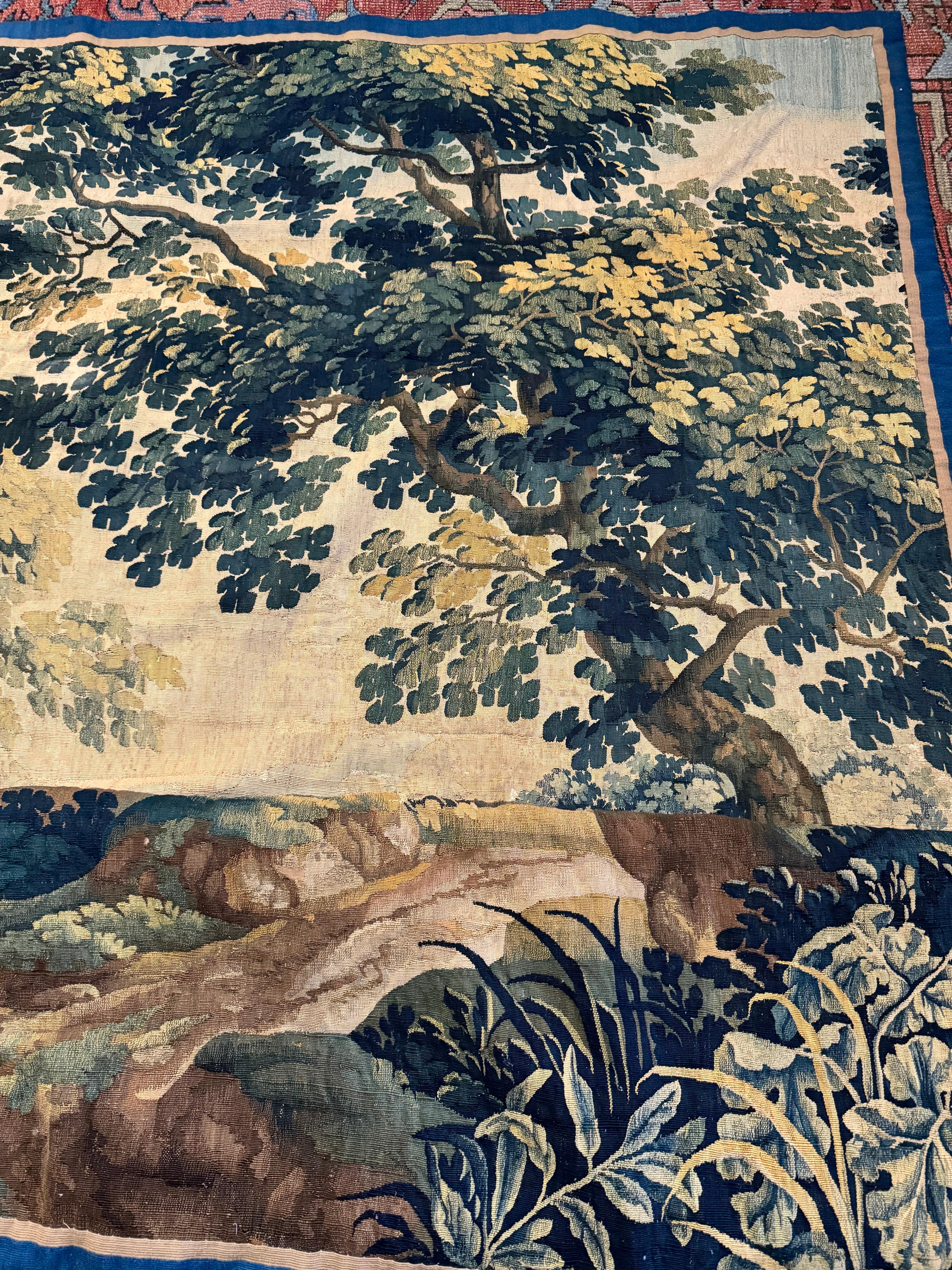 Se trata de un finísimo tapiz paisajístico cuadrado de Verdura flamenca de finales del siglo XVII con una bella y rica escena veraniega de una campiña con frondosos árboles y vegetación un castillo y una cascada a lo lejos

La pieza mide 2,5 x 2,5