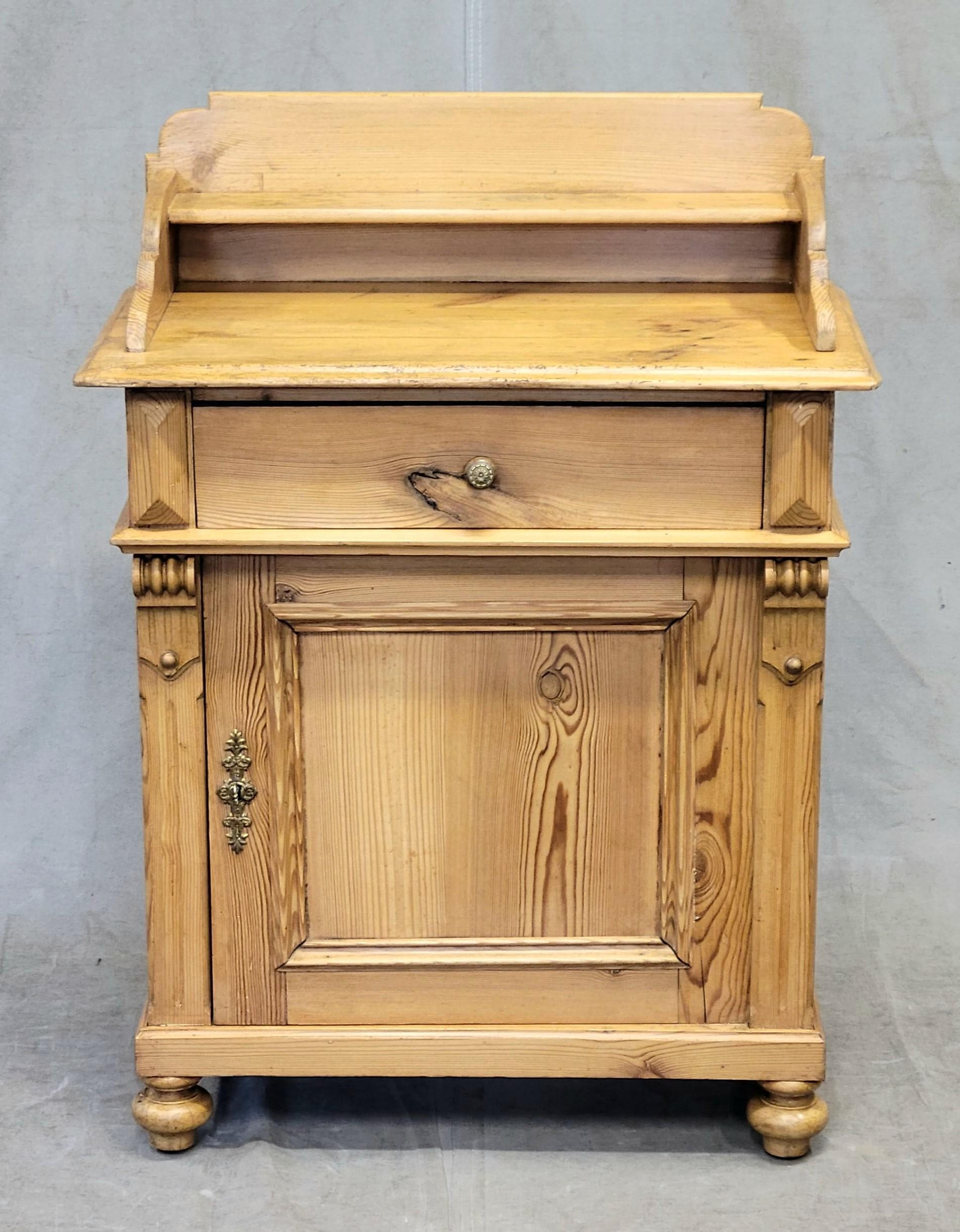 An antique sweet and petite late 1800s European, likely German, pine commode cabinet. Hand carved accents, including a crown with shelf, and distressed pine from age and wear give this piece such character. Old brass hardware is a wonderful accent.