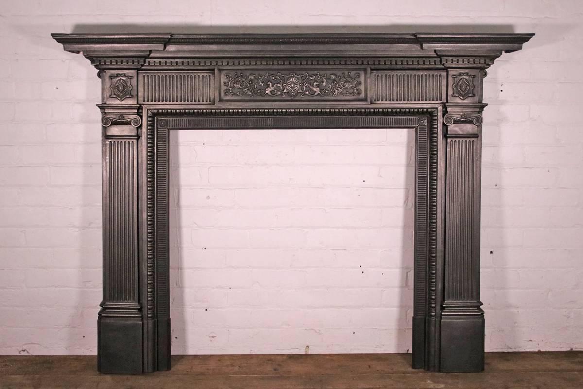 A substantial antique late 19th century Victorian cast iron fireplace surround. An inverted breakfront stepped shelf sits above dentil moulding and a fluted frieze with a detailed central panel of acanthus leaf scrolls, winged lions and a central