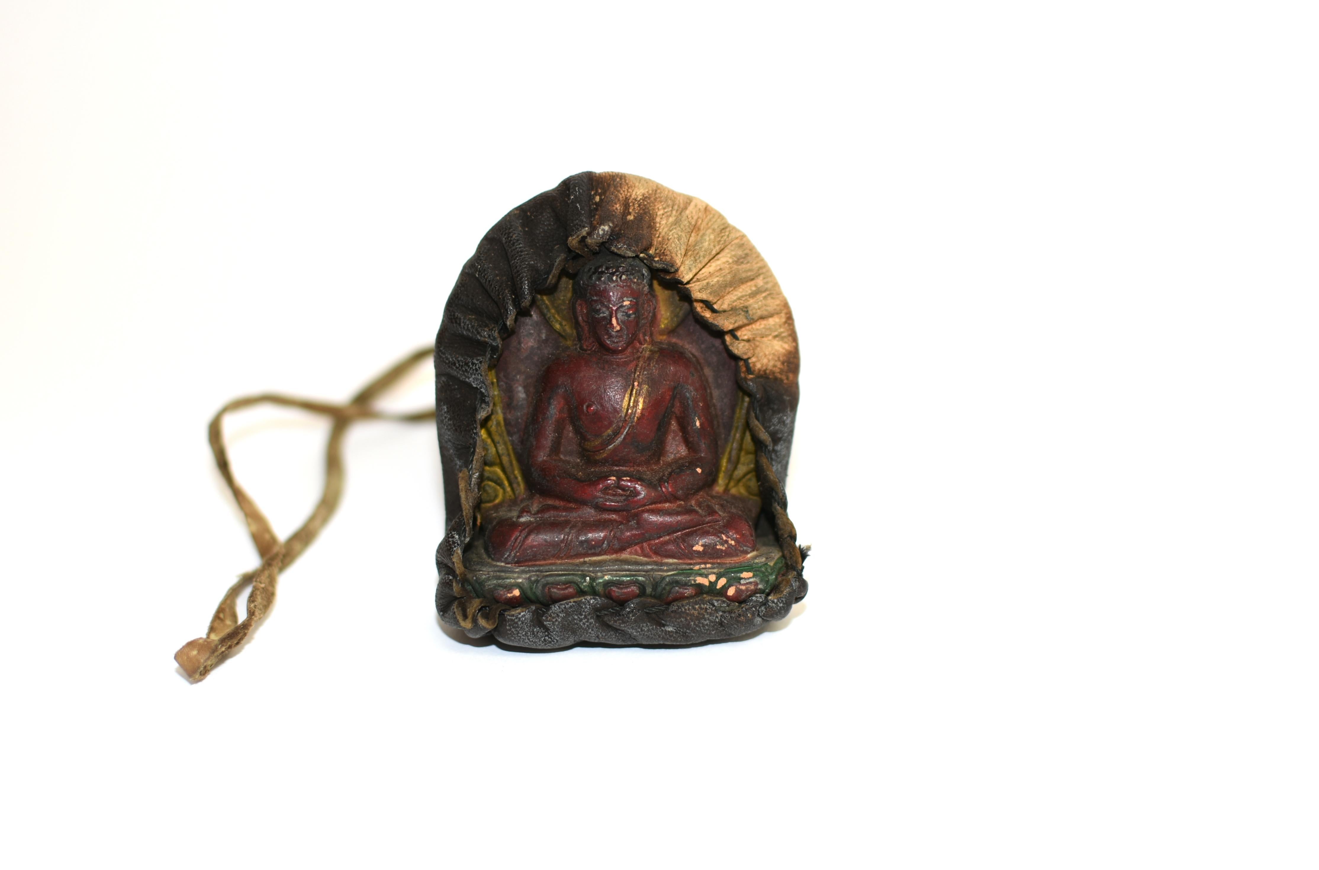 A Tibetan amulet featuring Buddha Shakyamuni encased in a hand stitched leather shrine. Seated dhyana asana on a lotus throne, with downcast eyes flanked by long earlobes under neatly coiled hair. He wears a robe that leaves his right chest bare and