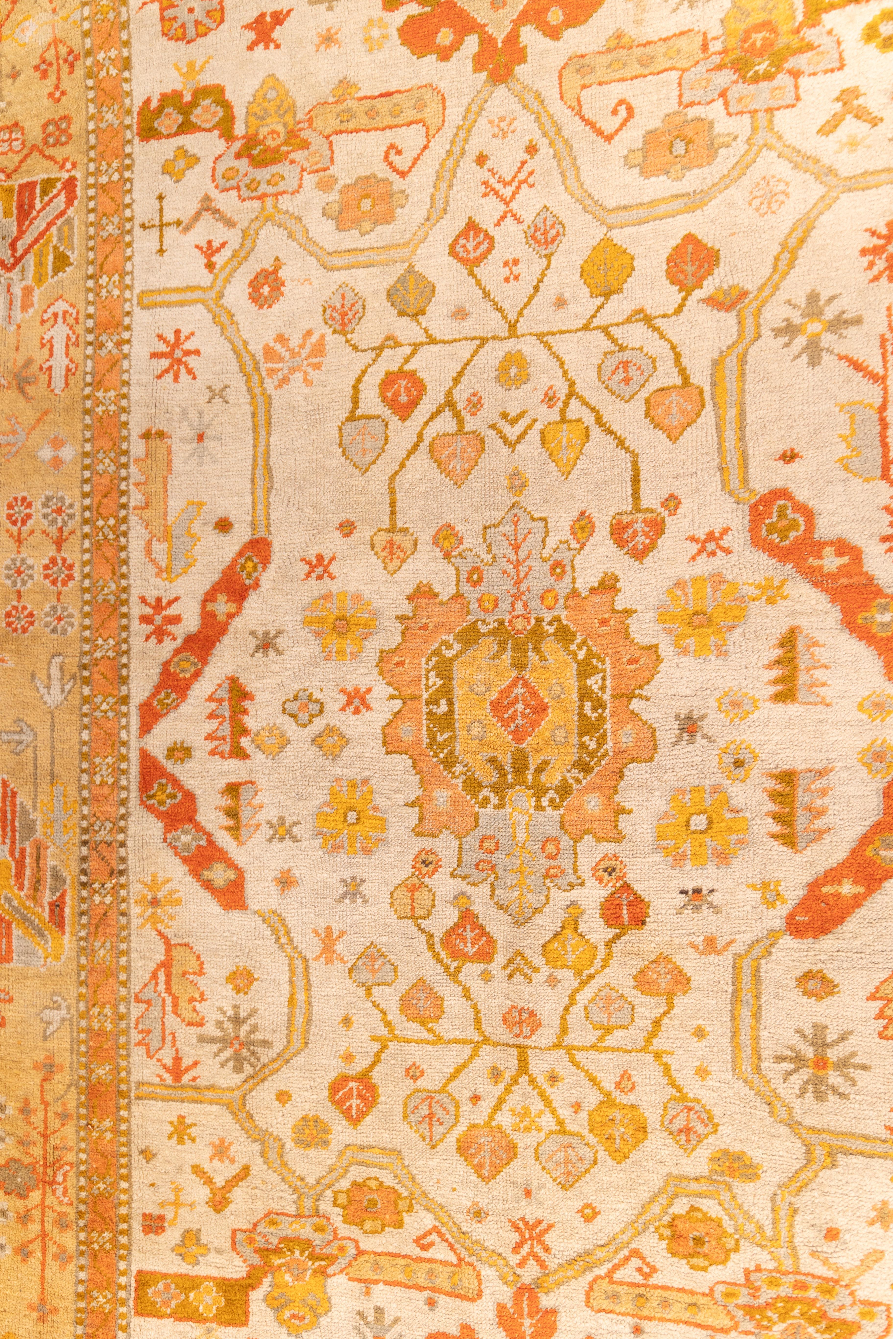 small antique Oushak circa 1910 featuring a soft ivory field with a bold central medallion in warm terracotta. The border blends gold, apricot, and sand tones with simple floral and vine motifs. Light scattered geometric designs give it a relaxed,