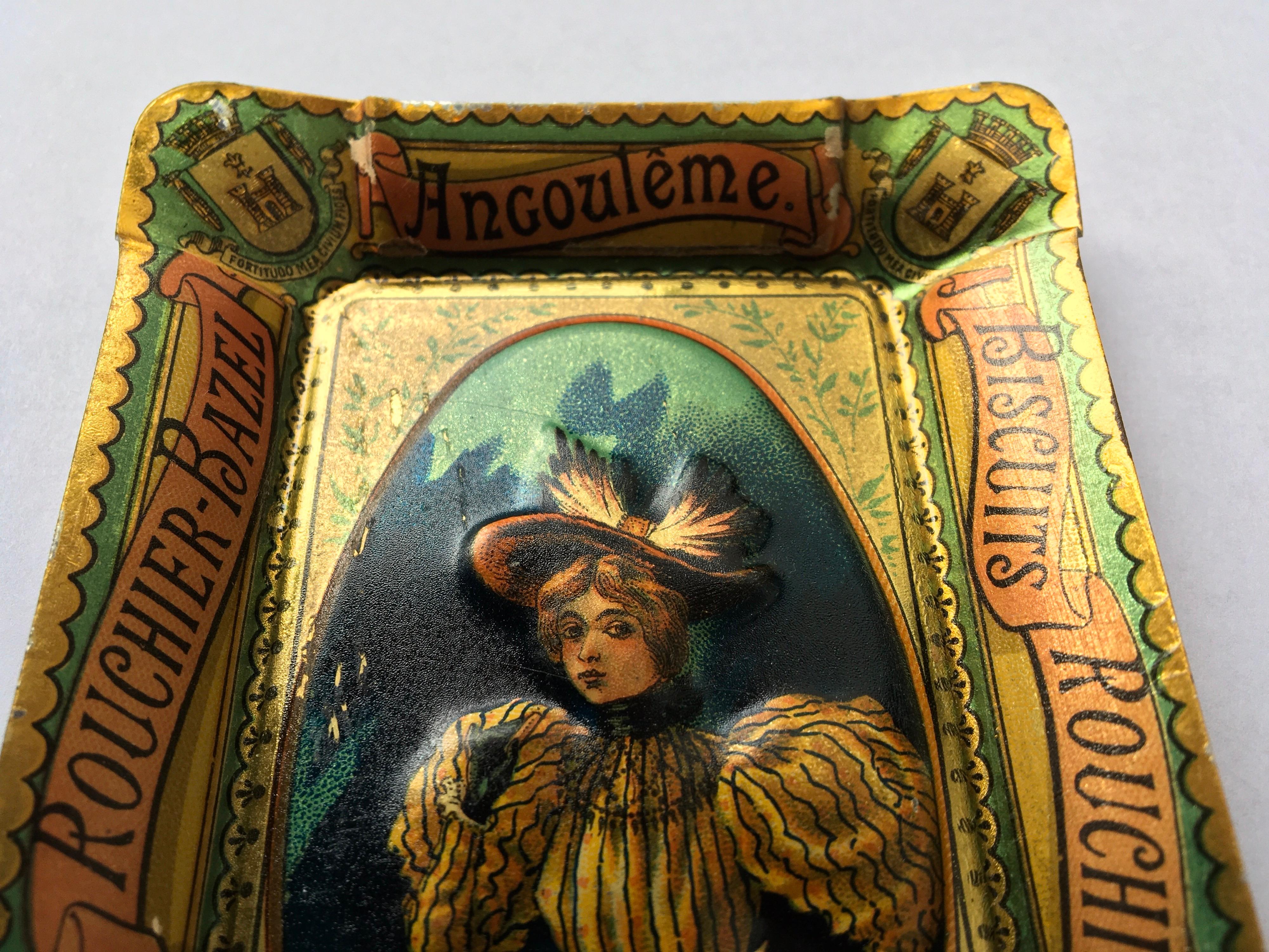 Antique Tin tray, dish or ashtray for Biscuits Rouchier Bazel wich was located in Angoulême France. 
This French lithographic and embossed tray dates circa 1900. It shows an elegant lady holding a box of biscuits and wearing the typical clothing of