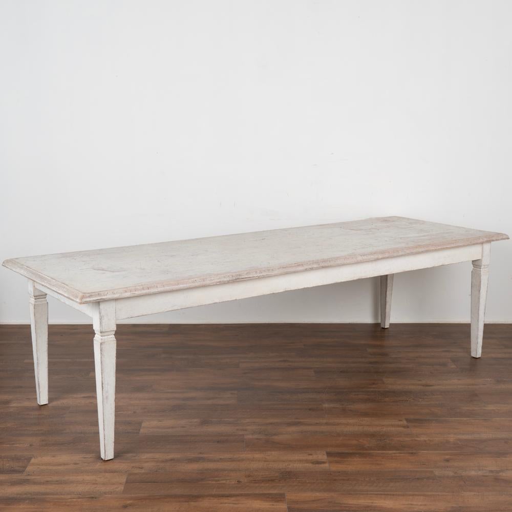 Long antique white painted dining table with four tapered legs reflecting its Swedish country styling.
The white paint has been softly distressed over years of use, revealing the warm patina of pine wood below. 
Restored, strong and stable this