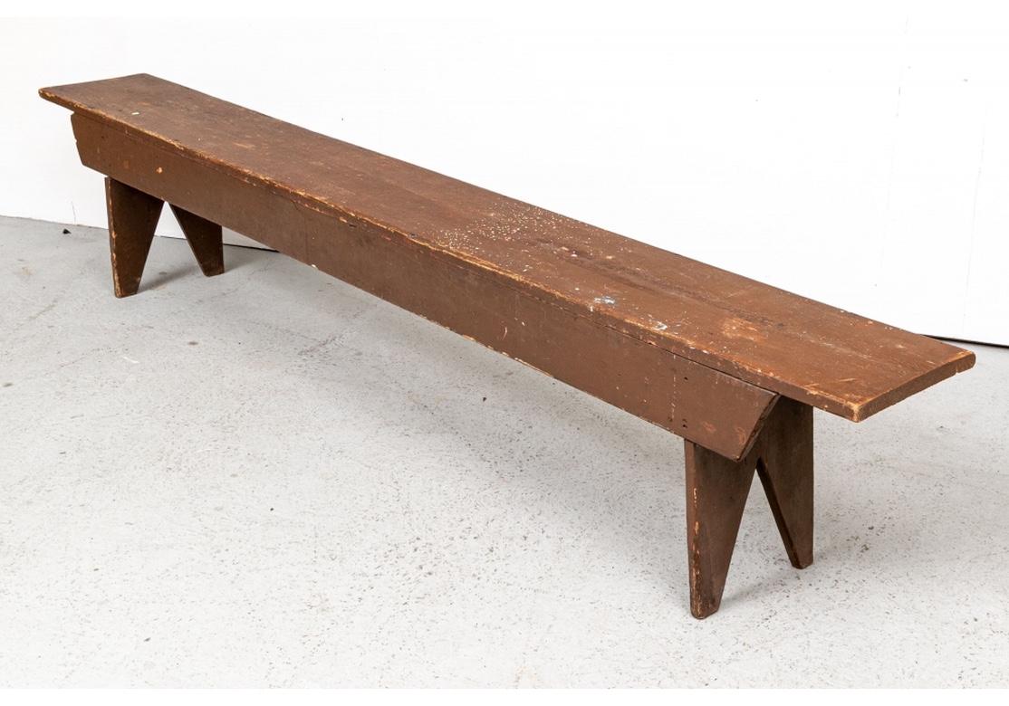An antique wooden bench in old brown paint with other paint spatters here and there on top. The extended top with attached sides is mounted on notched carved flat legs on each end. An authentic and solid Antique Bench in all original condition.