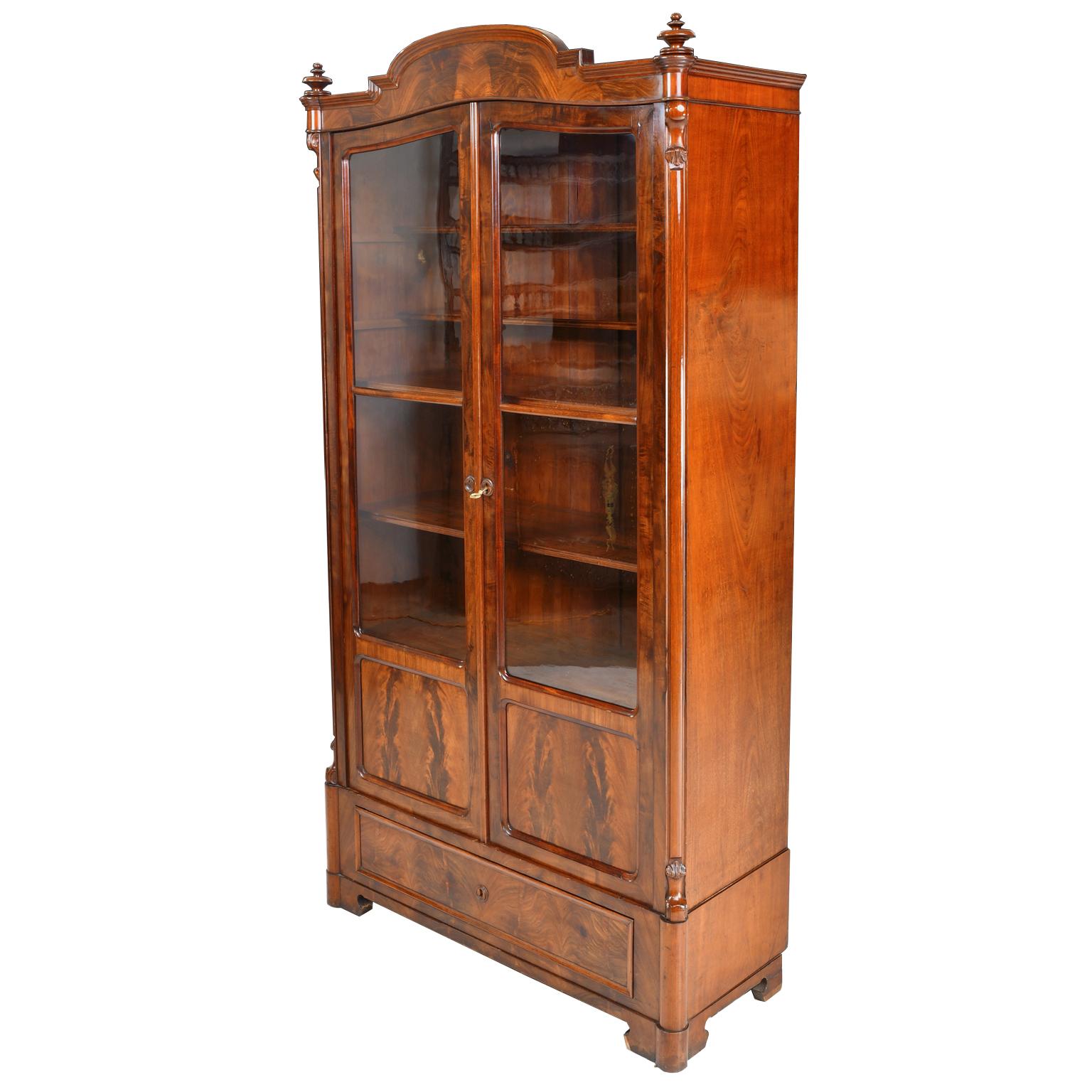 A beautiful Louis Philippe bookcase in fine Cuban/ West Indies mahogany with arched bonnet top over two doors, with original glass panels, that open to adjustable shelving. Cabinet offers one exterior drawer along the bottom and rests on original