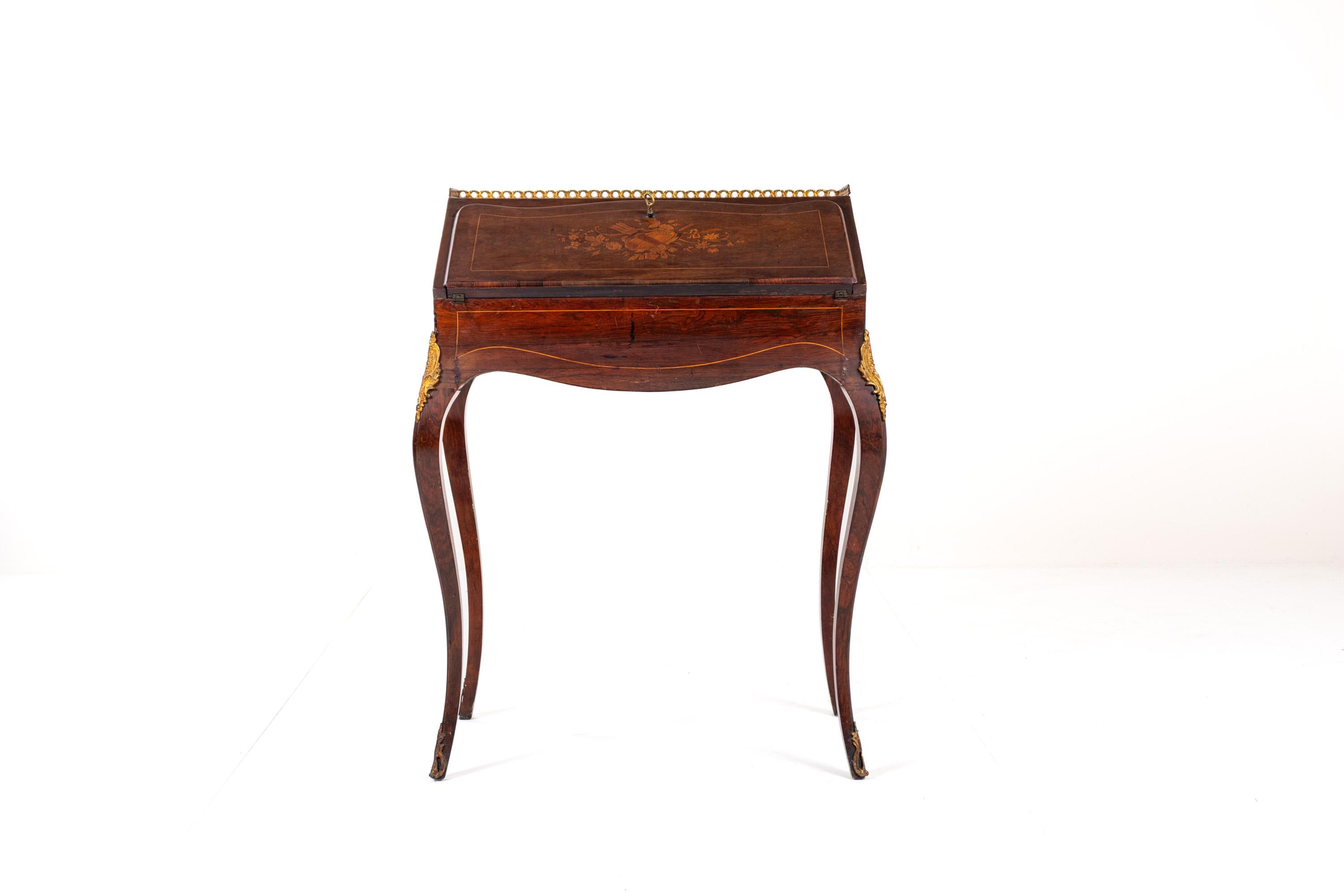 Exquis bureau ancien de style Louis XV, en placage de bois de rose avec incrustations de marqueterie et orné de montures en bronze doré (ormolu).
La silhouette serpentine, les pieds cabriole élancés et la galerie en laiton sur le plateau définissent