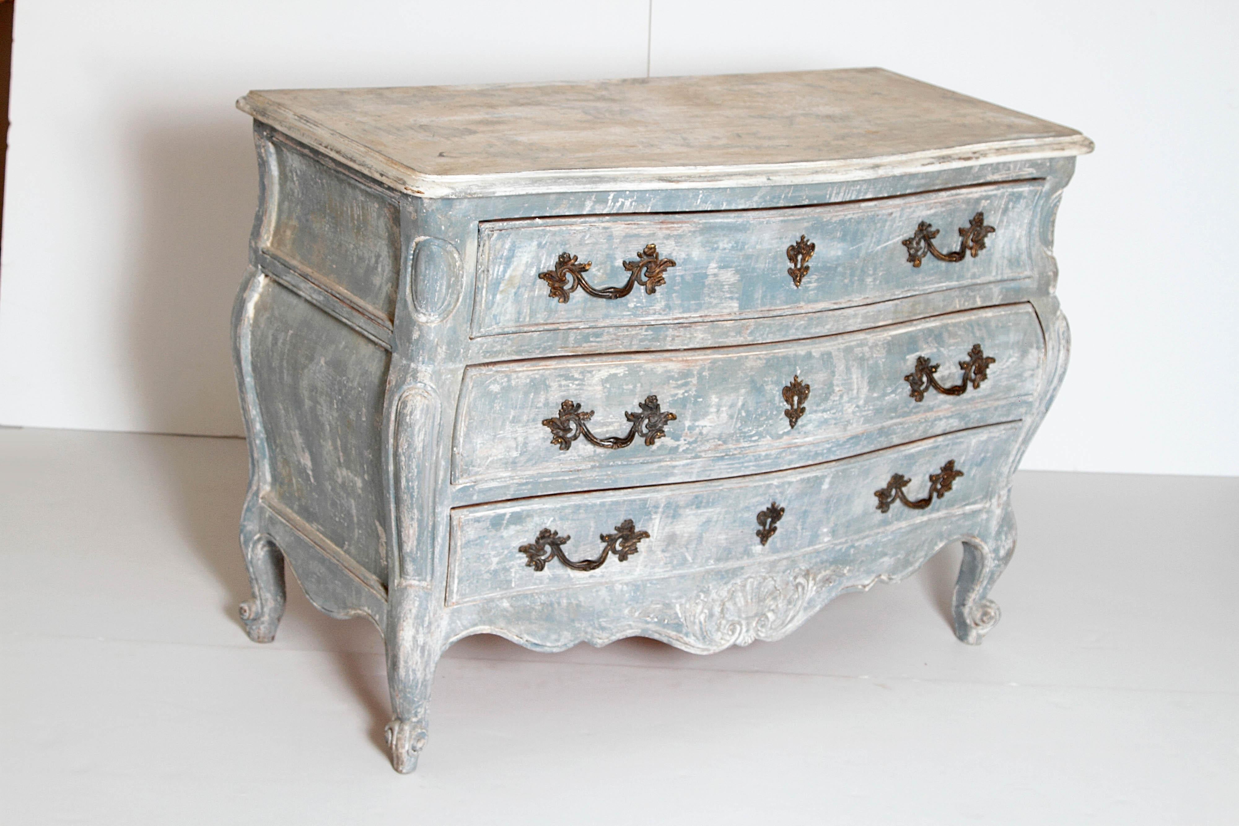 This is a wonderful antique French commode with a weathered painted finish and a serpentine wood top. The carved bombe shape sides terminate in cabriole legs with scroll feet. The three drawers with original bronze pulls and key hole escutcheons sit