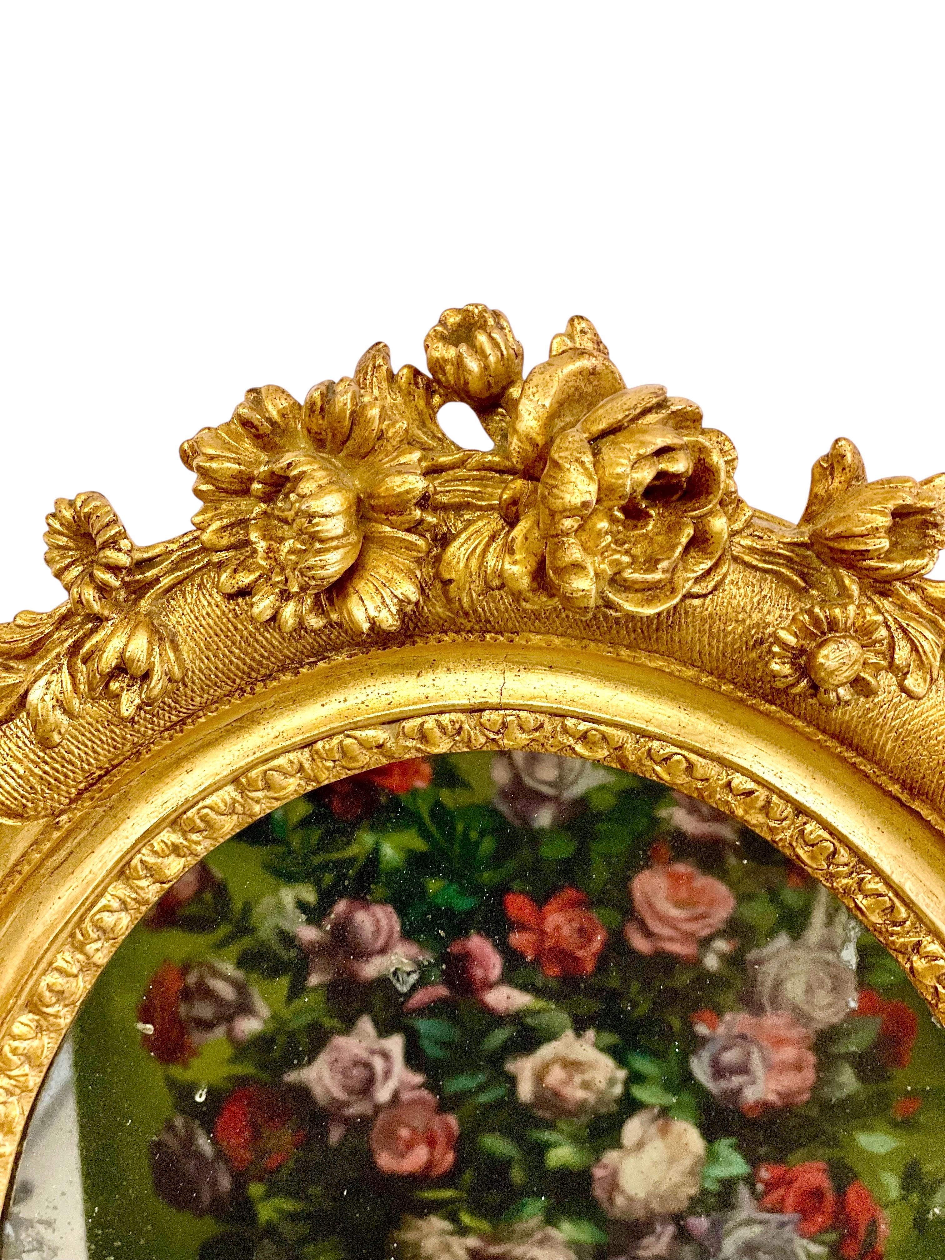 This exquisite and romantic 19th century French oval mirror features an elaborately carved and gilded wooden frame, richly adorned with finely detailed floral motifs in high relief. Crafted in the Louis XVI or possibly late Louis XV style, the frame