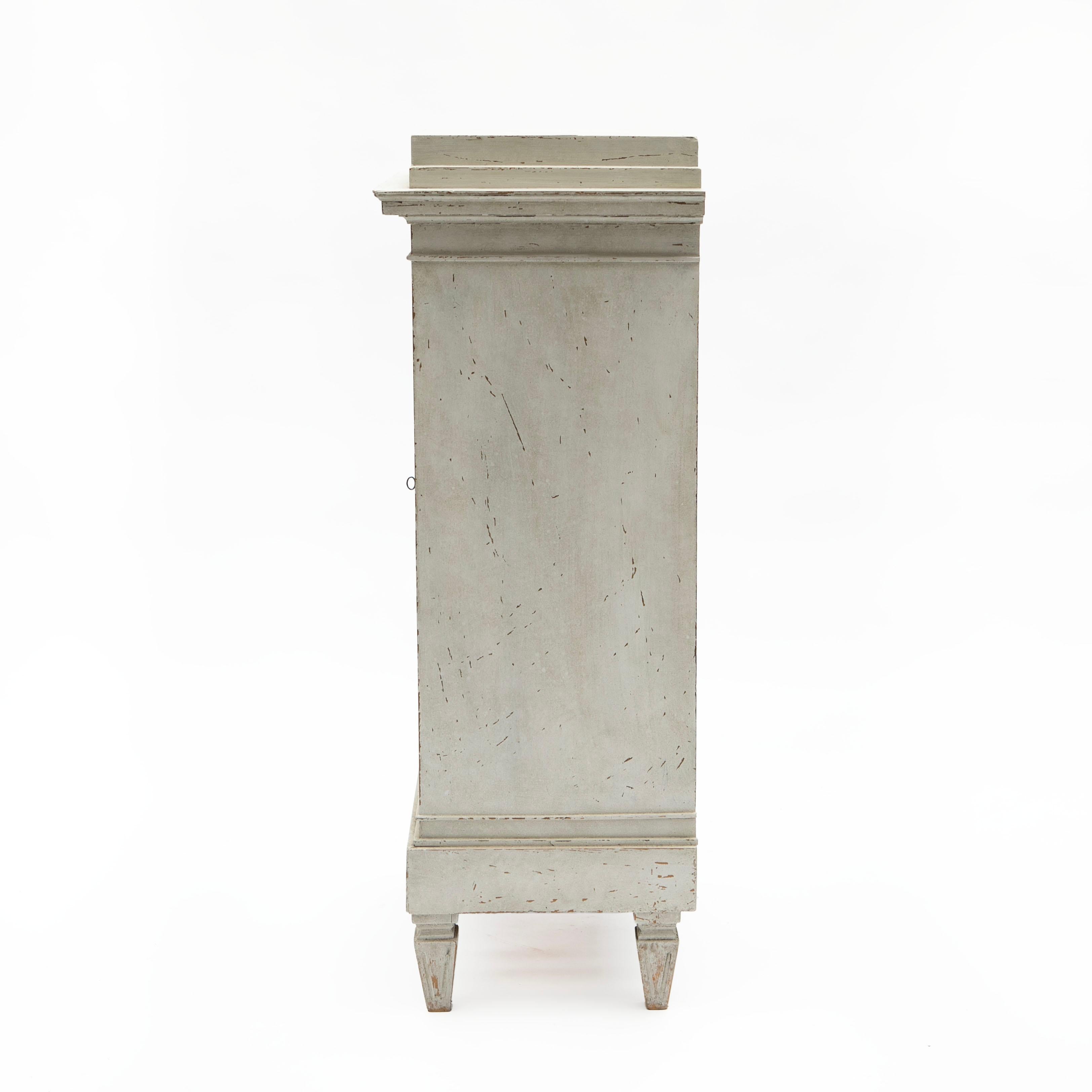 Louis XVI pedestal cabinet in a beautiful gray color.
Features a door with a wooden-carved sun in the center flanked by flutings on each side. At the top, there's a wooden-carved Vitruvian scroll also known as 'running dog' pattern. Resting on four