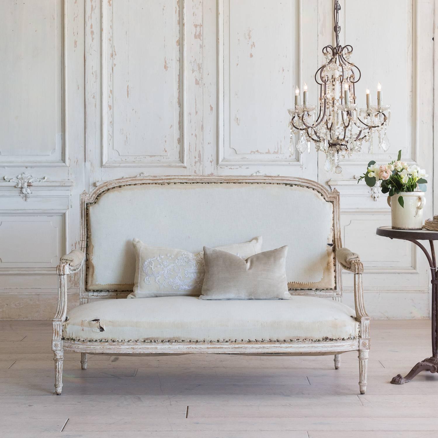 Adorable antique loveseat in distressed cream with lots of raw wood peaking through the finish. The Classic Louis XVI style boasts simple, grooves along the frame and small, architectural finials adorn the shoulders of this sofa. Shown in original