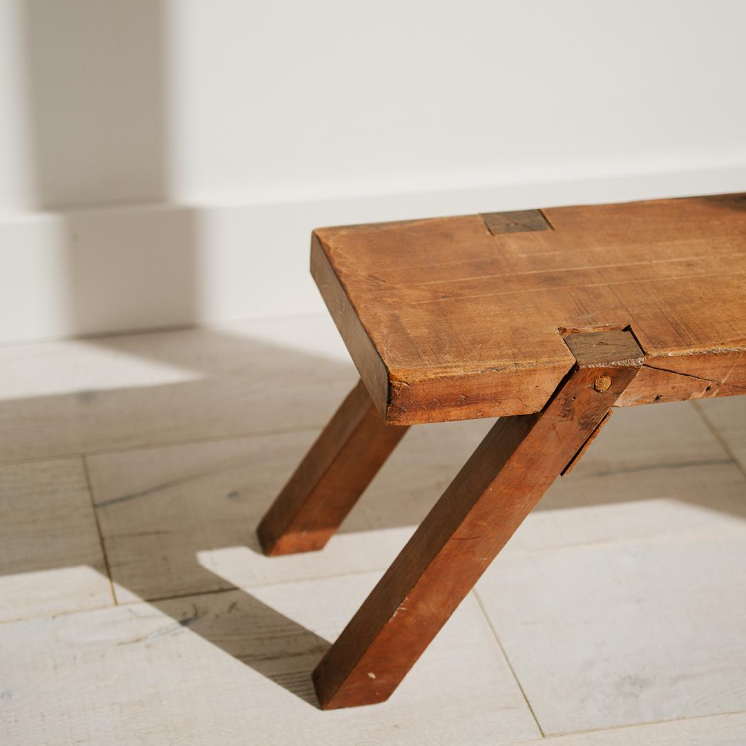 Ce banc bas antique présente de charmants pieds en épingle et une surface en bois magnifiquement vieillie, mettant en valeur son caractère rustique et son savoir-faire artisanal. Son design simple et discret ajoute de la chaleur et un sens de