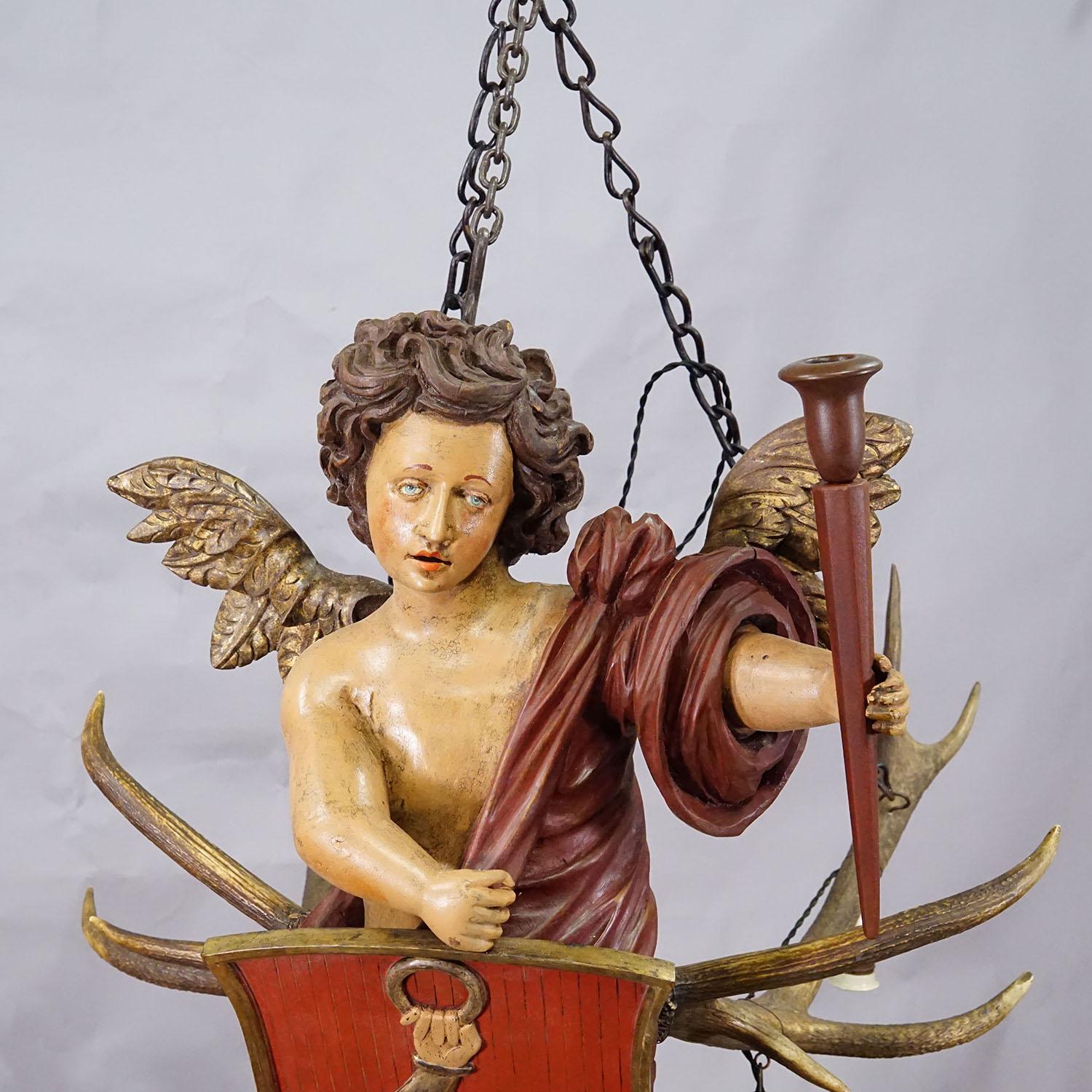 Antico Angelo Lusterweibchen, Germania 1900

Un raro Lusterweibchen storico a forma di putto alato, finemente intagliato e dipinto in policromia. La figura, avvolta in una veste rosso bordeaux, regge uno scudo araldico con un braccio eretto che