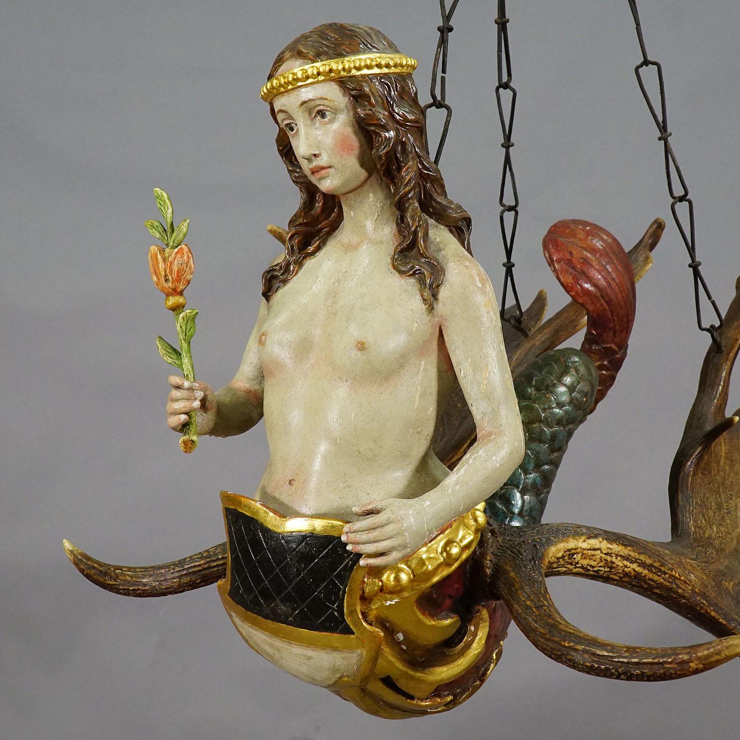 A wonderful antique lüsterweibchen with a carved wooden sculpture of a mermaid with fishtail. The maid is holding a flower and a heraldic blazon. Mounted on the sculpture are a pair of original fallow deer antlers and a hand forged iron suspension.