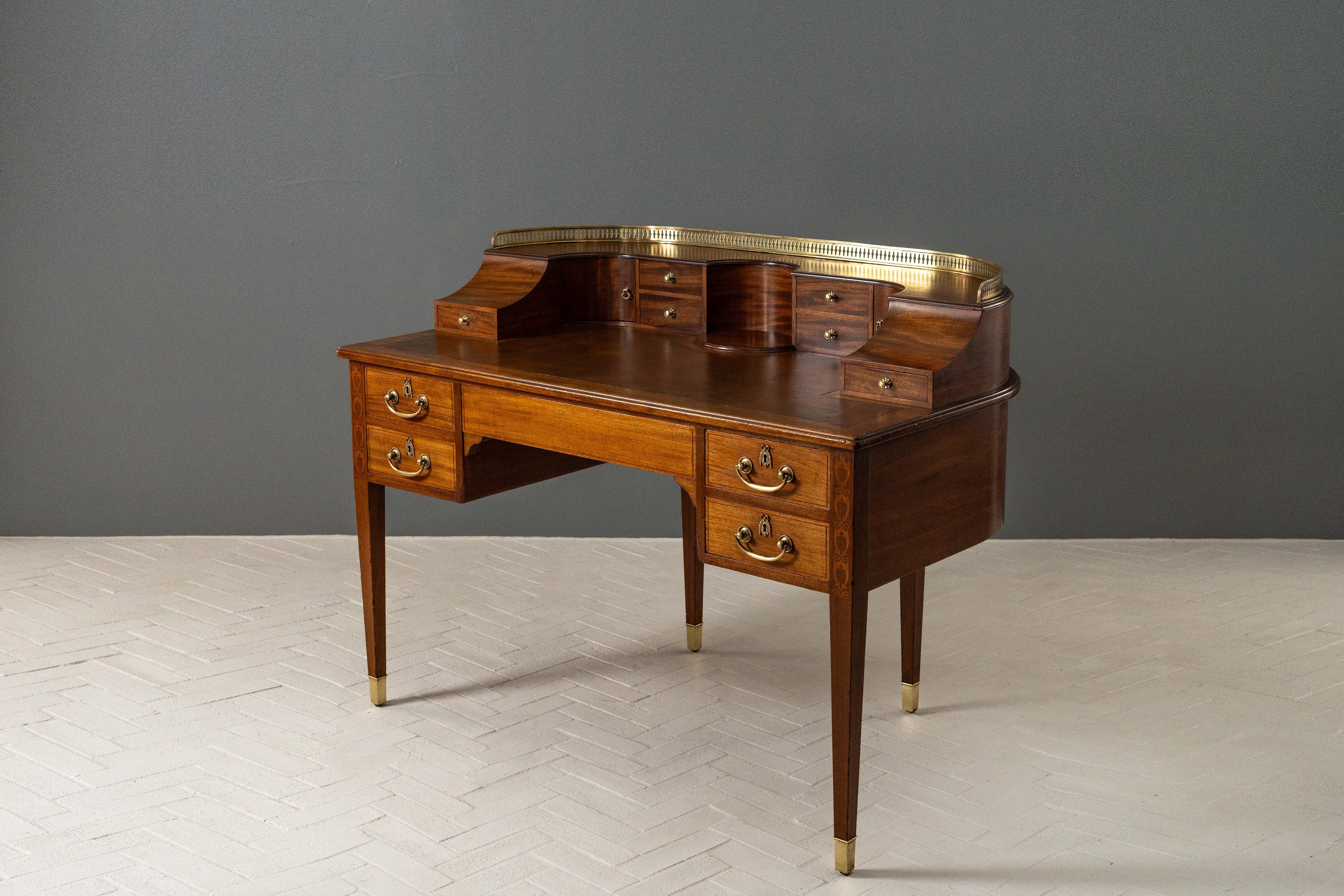 England, ca. late 19th century. An elegant Carlton House desk after an earlier design by George Hepplewhite. Features mahogany construction with subtle satinwood inlay and inset brown leather writing surface. Lower portion includes five drawers -