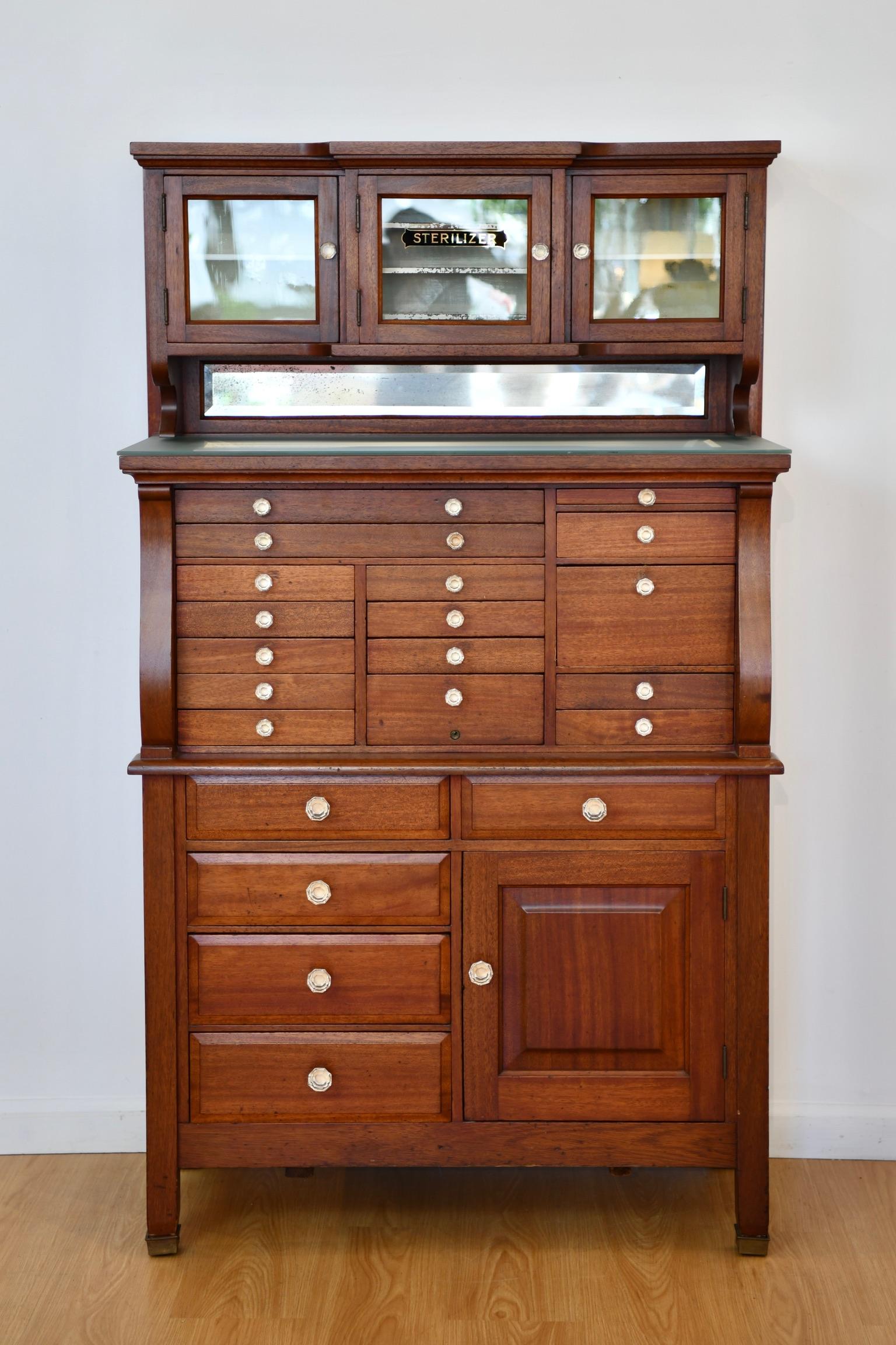 Antique mahogany dental cabinet fitted with twenty-one drawers, four cupboards, and frosted glass top, by American Cabinet Co and circa early 20th century. Dimensions: 61.25