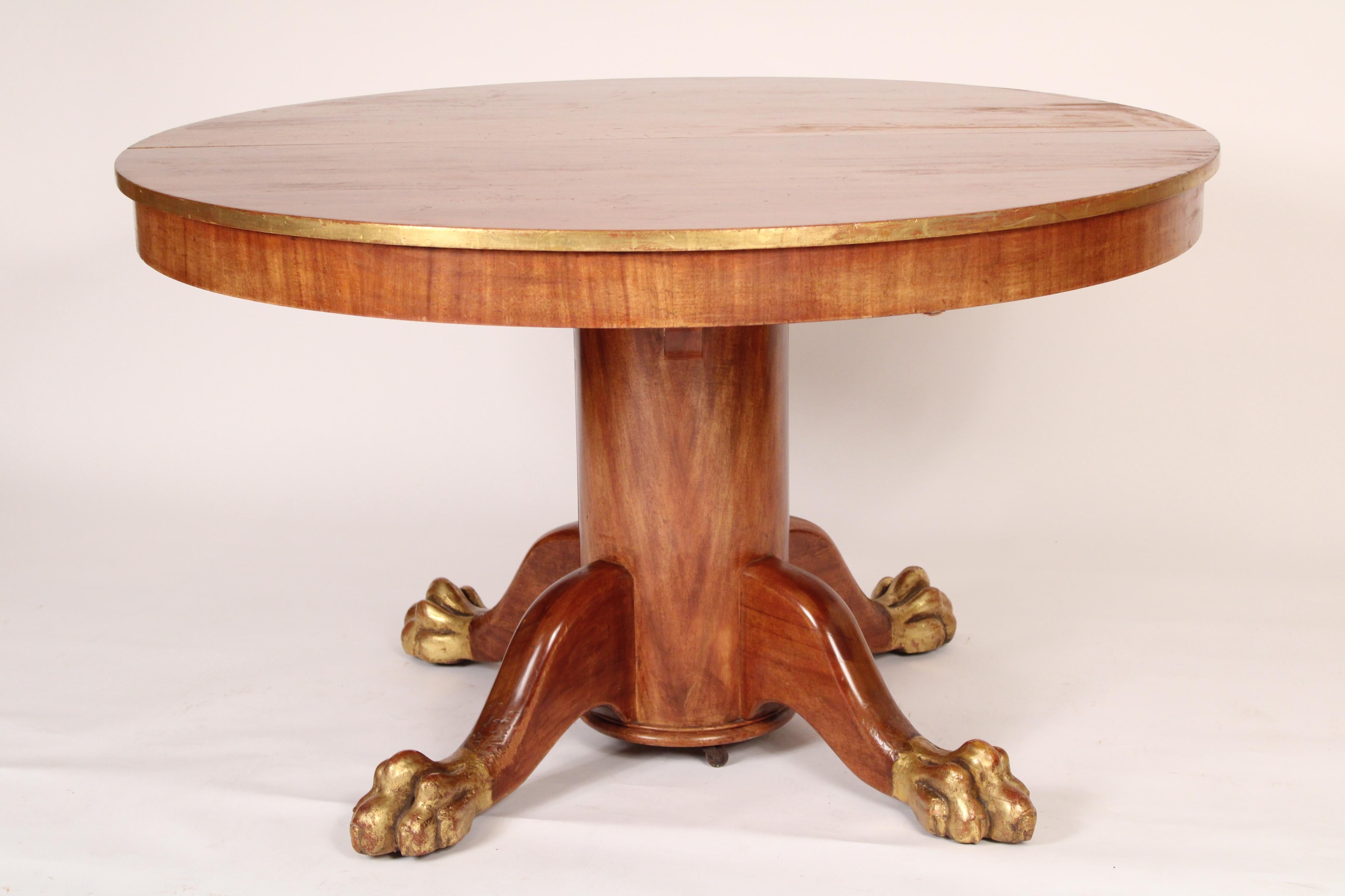 Ancienne table ronde de salle à manger de style Empire en acajou avec des reflets dorés, vers 1900. Avec un plateau rond qui s'ouvre (pas de feuilles), une décoration dorée sur le bord du plateau, un piédestal central rond, quatre pieds se terminant
