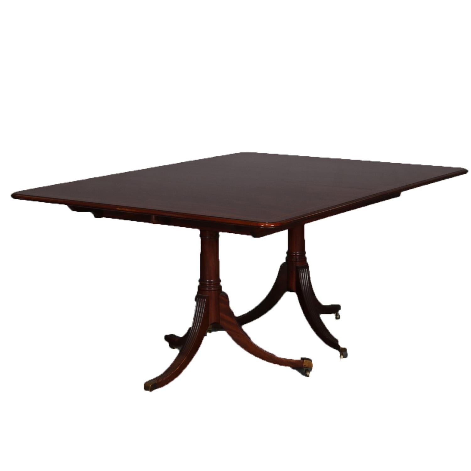 Antique Duncan Phyfe School dining table features mahogany construction with cross banded top having satinwood inlay, single leaf and raised on double tripod pedestals each having three reeded concave legs terminating in bronze caps with casters,