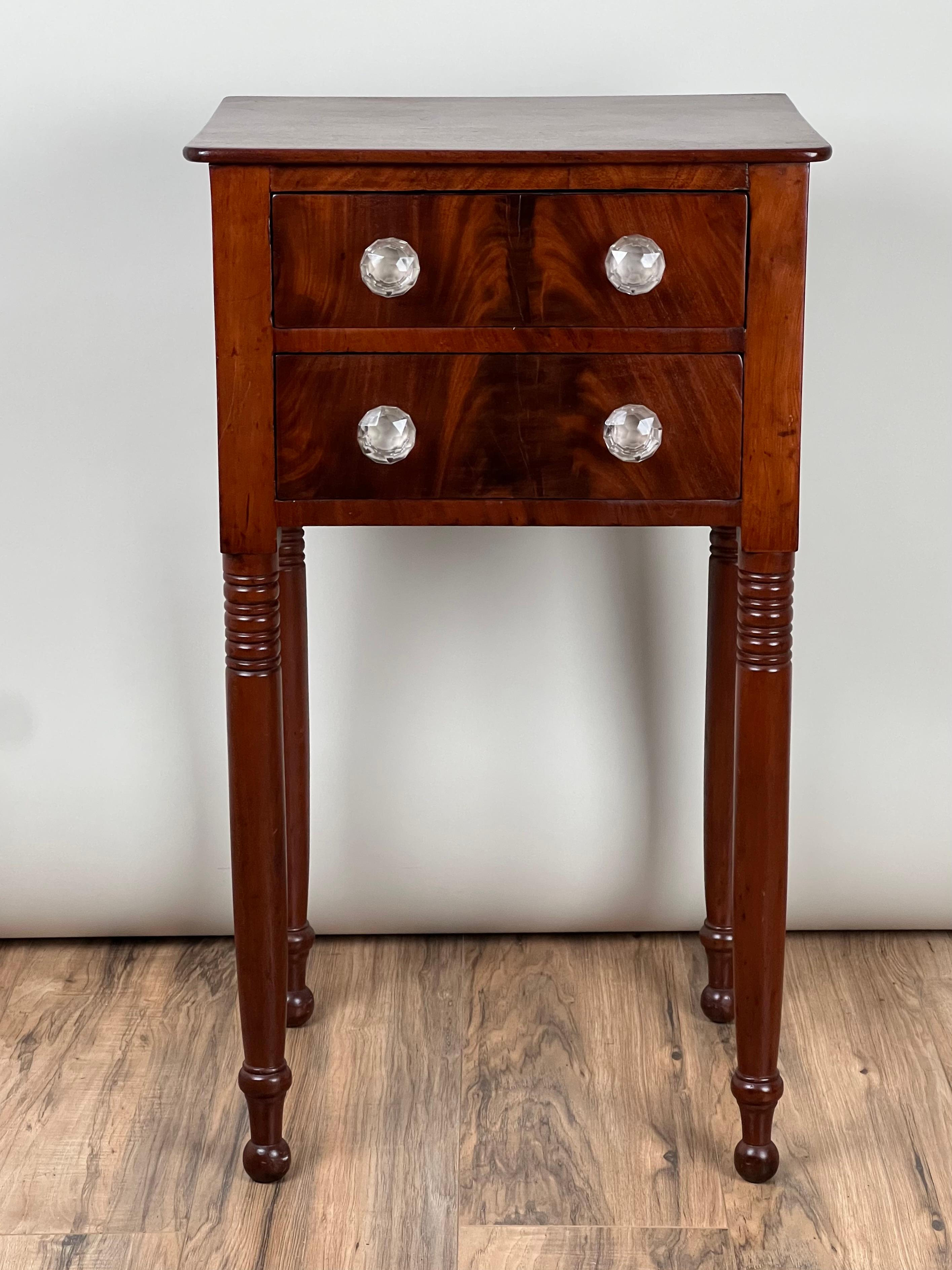 Ce charmant meuble à deux tiroirs, datant d'environ 1820, illustre l'élégance du design de la période fédérale, en particulier du style Sheraton. Fabriquée en acajou, cette pièce met en valeur la chaleur et la profondeur du grain naturel du bois. Le