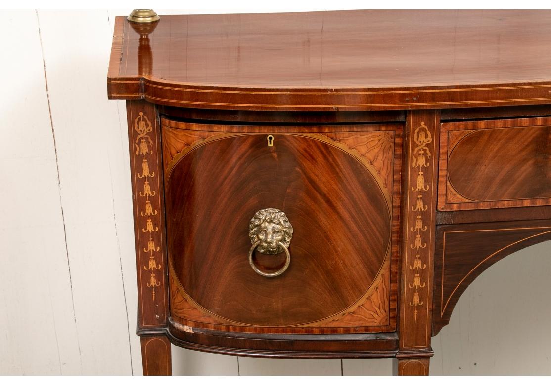 The cabinet with a shaped bow front top with satinwood banding and a fine brass open back support with urn form finials. With a long center burled apron drawer with satinwood banding and corner inlays with quarter sun rays. The knee hole opening