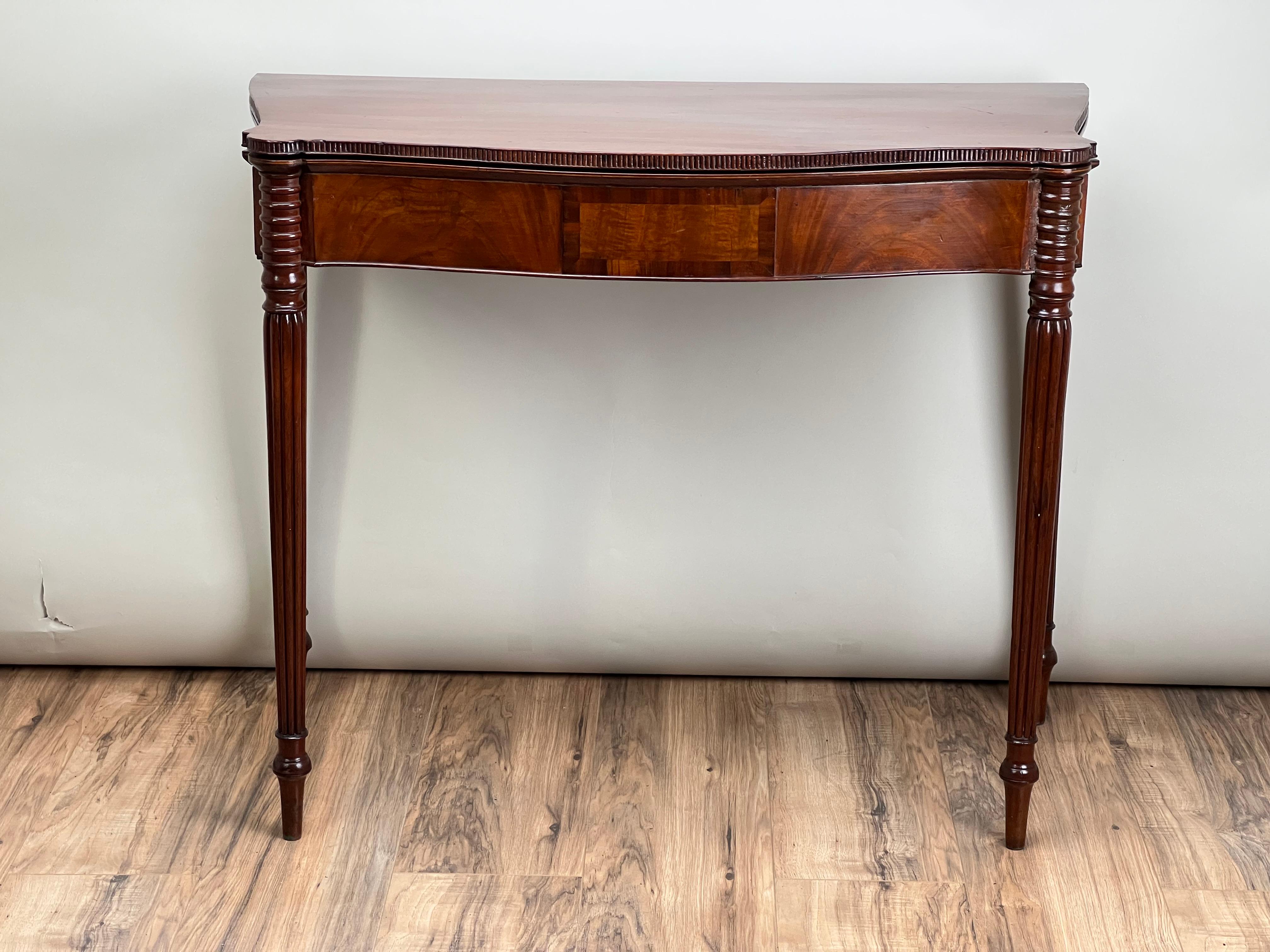 Table de jeu Sheraton du début du XIXe siècle, fabriquée en acajou riche, aux proportions élégantes, avec une façade serpentine qui met en valeur sa silhouette gracieuse. Le plateau rabattable révèle une surface de jeu spacieuse, soutenue par un