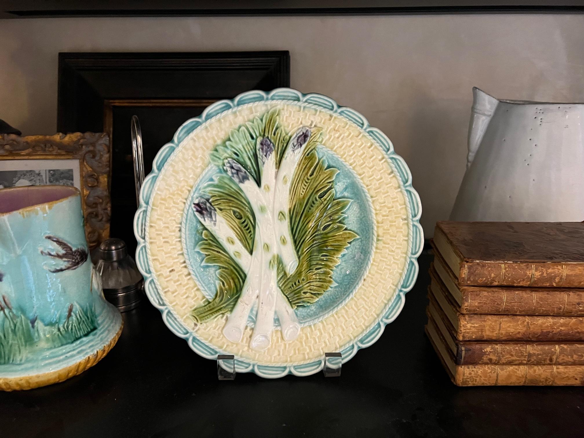 A French majolica asparagus plate from the Salins-les-Bains region of France, made in the late 19th century. The plate has four white asparagus with purple tips, laying on a bed of green leaves. The plate includes a sauce well and is numbered on the