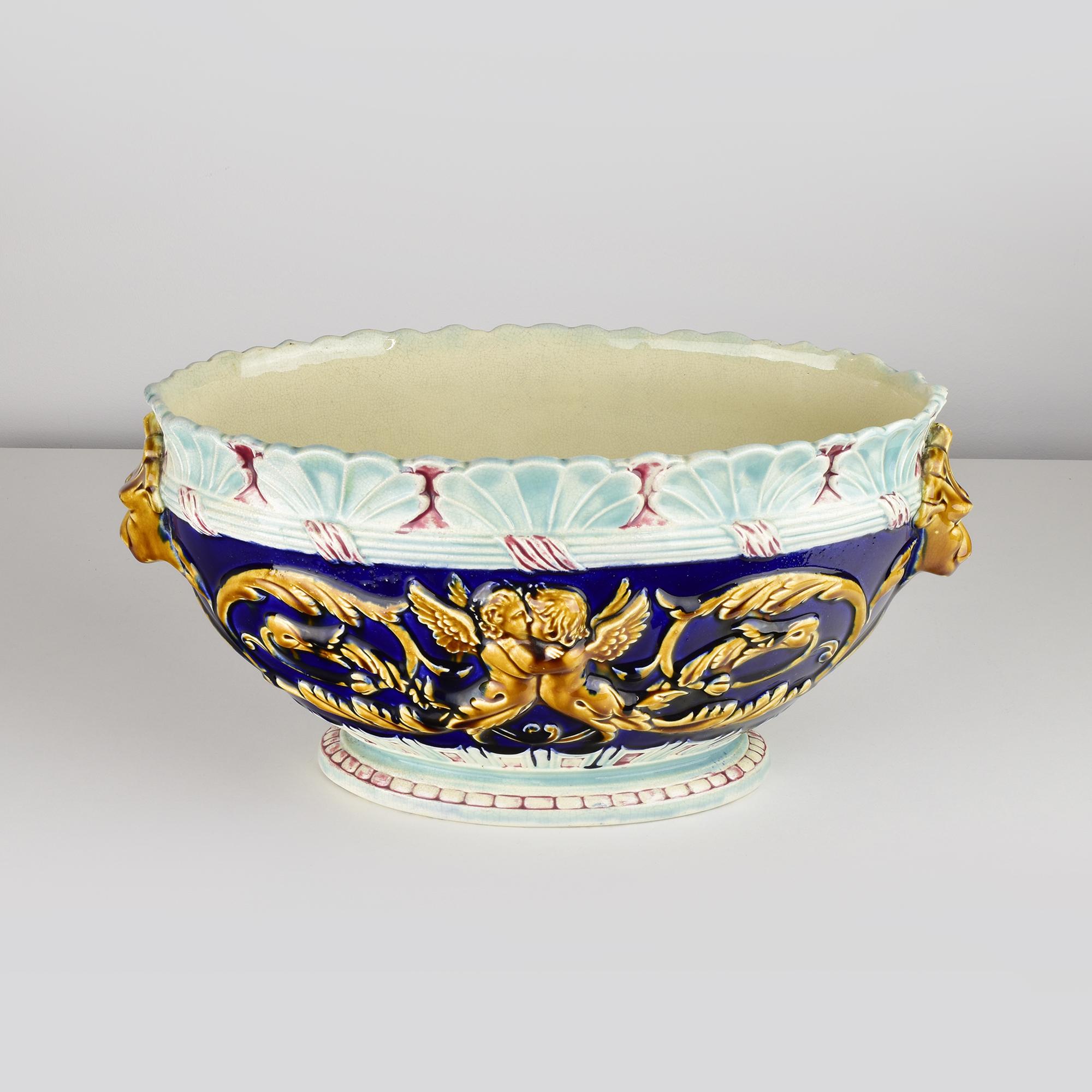 This Majolica jardinière is richly decorated, featuring a deep cobalt blue glaze as the base color. The sides are adorned with cherubs and lion heads in golden and brown tones, reflecting a classical or baroque style. The rim of the jardinière