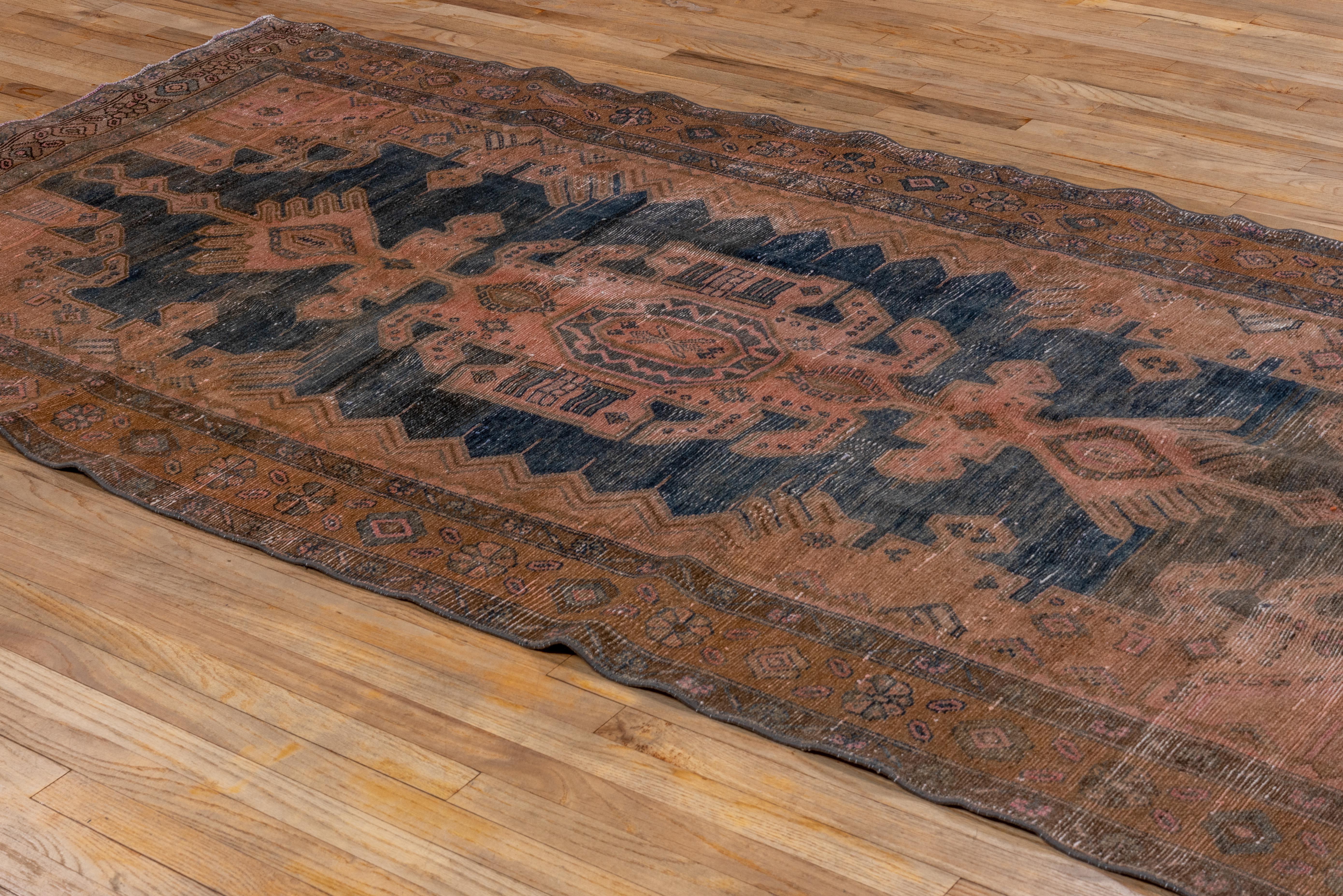 This antique Malayer rug presents a dramatic central medallion in inky navy with stepped and angular detailing, set against a weathered terracotta ground. The field features elongated botanical forms and stylized spandrels, framed by a softly worn