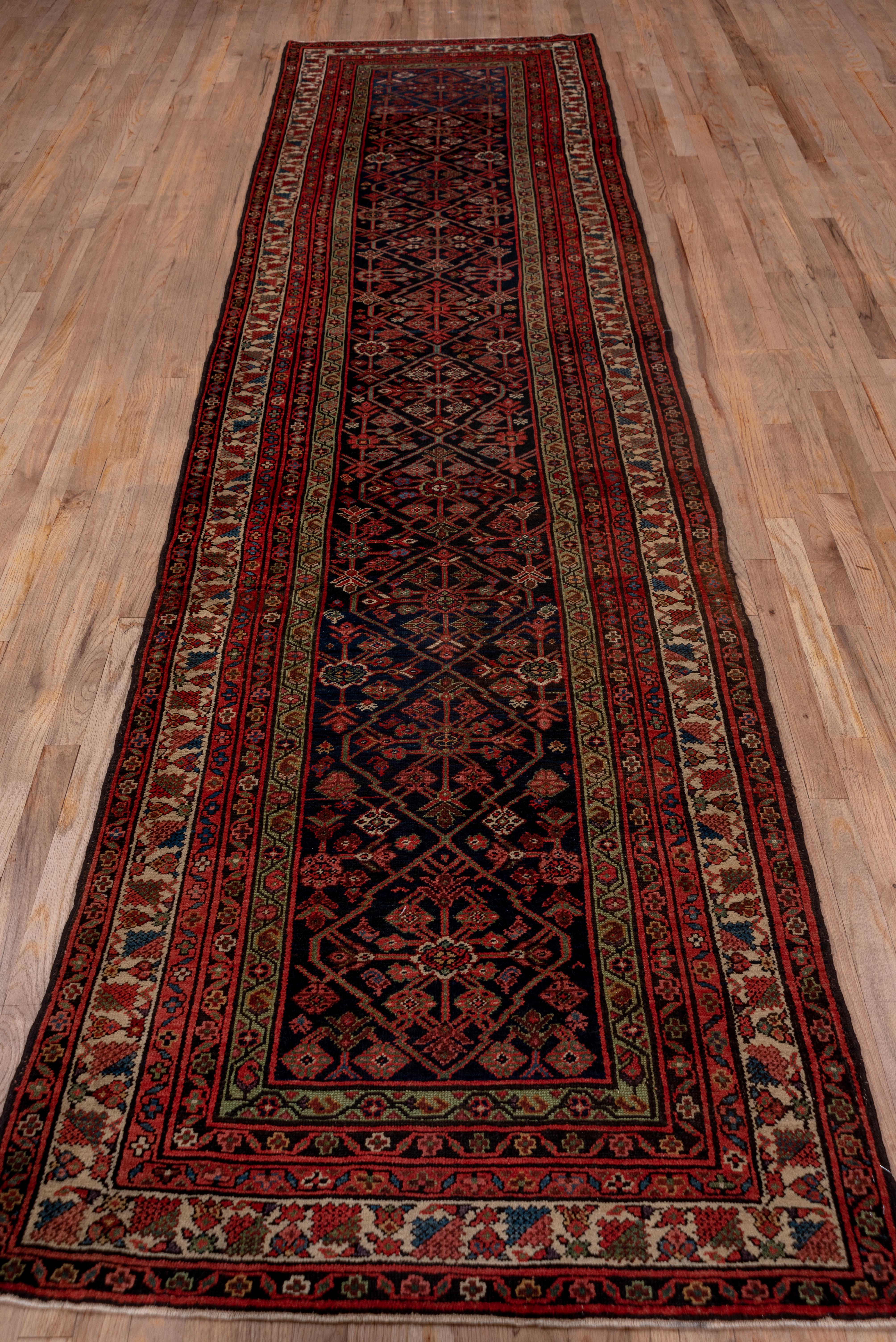 This west Persian village runner shows a navy ground supporting a skeletal connected lozenge pattern with central ivory rosettes. The ecru main border features square palmettes and standing small flowers.