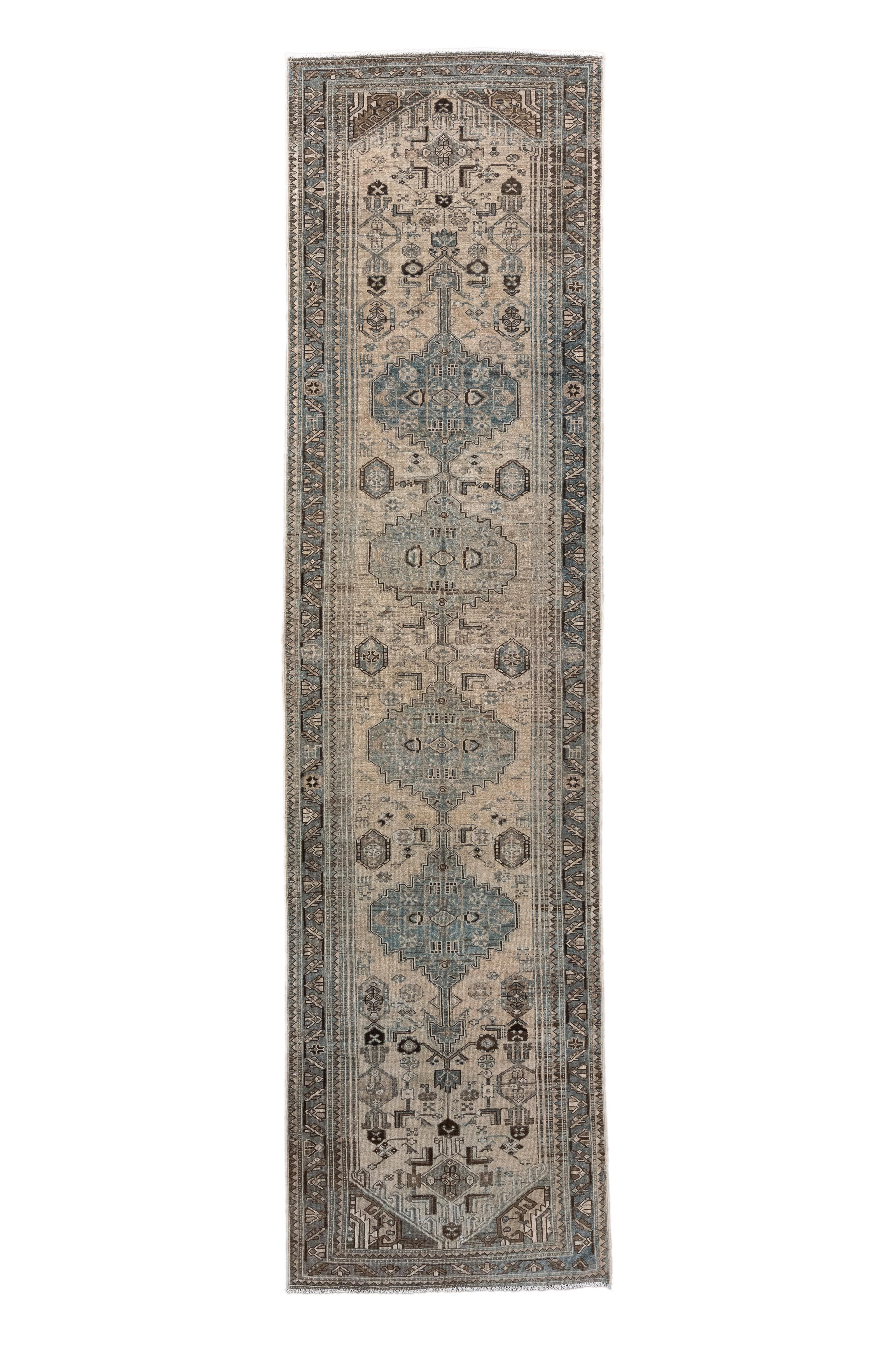  This antique Malayer runner, handwoven around 1910, features an elegant procession of stepped medallions in faded blue, ivory, and espresso brown, set within a soft oatmeal ground. The design is anchored by architectural corner spandrels and highly