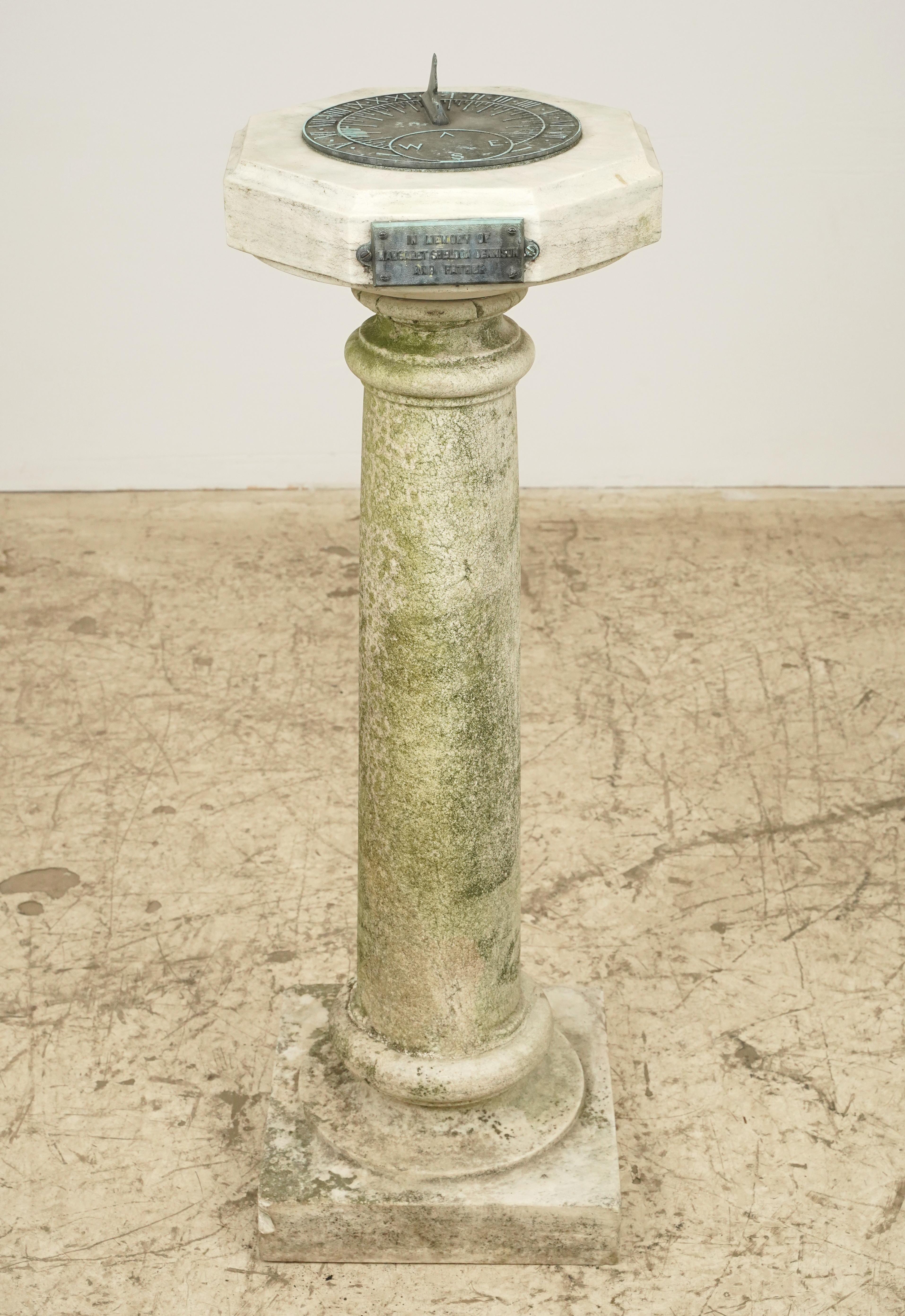 Antique marble sundial featuring a tall cylindrical pillar with a square base and top. The top section is a horizontal disk with a smaller bronze raised dial plate. The pillar is weathered, with moss growth, indicating age and exposure to the