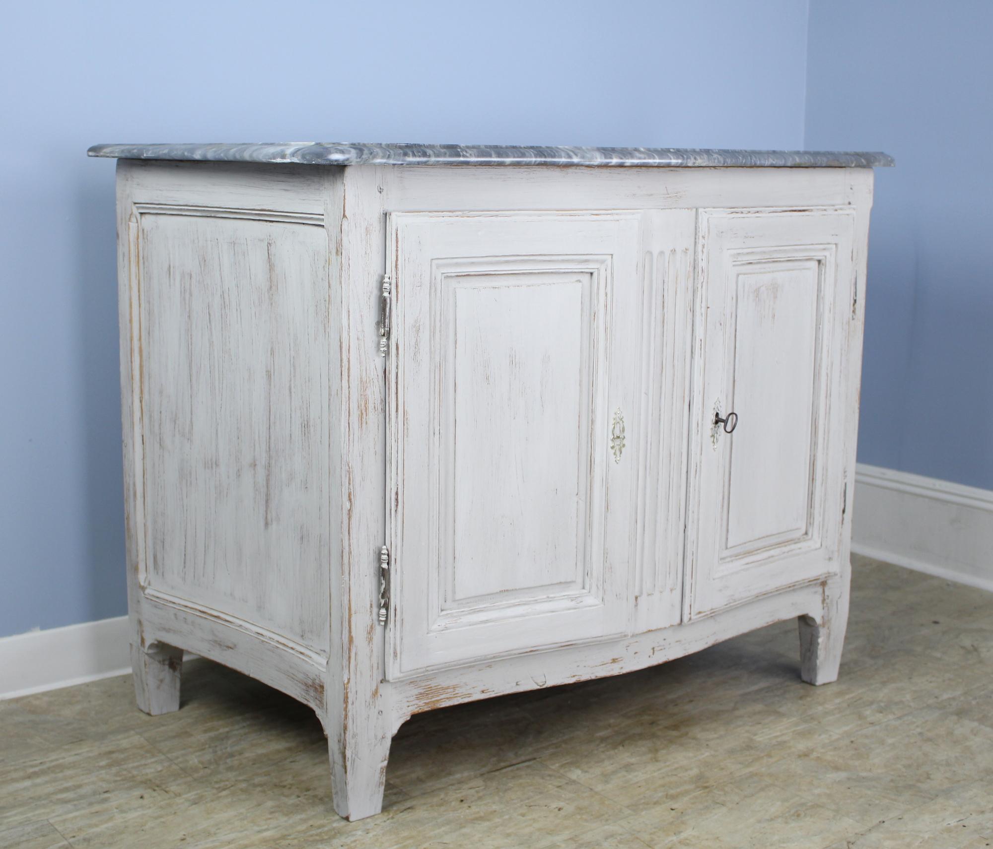 A handsome bowfront buffet, with original bright gray marble and newly painted in faux distressed white. The single interior shelf is not adjustable. The piece closes securely with an interior latch and lock and key. Marble is in good condition for
