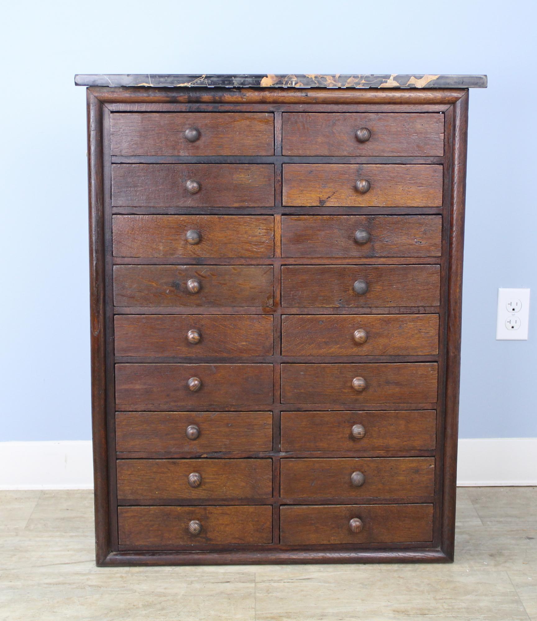 A handsome bank of drawers in dark brown oak. Marble top is dramatically grained and vibrant, with a small area of repair and some scratches. Each of the eighteen drawers is roomy and slides easily. There are no stoppers so the drawers are