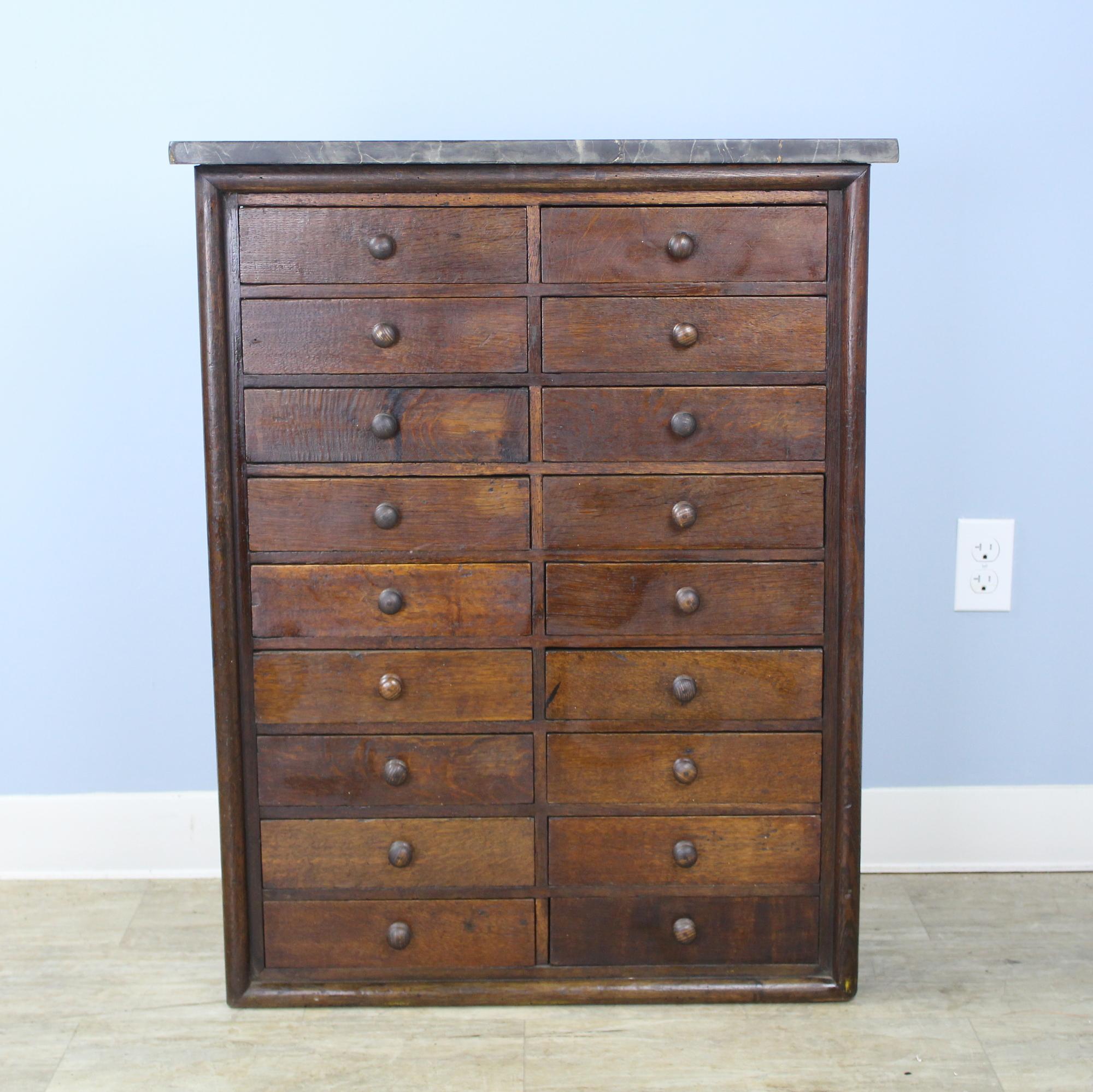 A handsome bank of drawers in dark brown oak. Marble top is dramatically grained and vibrant, with a small area of repair and some scratches. Each of the eighteen drawers is roomy and slides easily. There are no stoppers so the drawers are