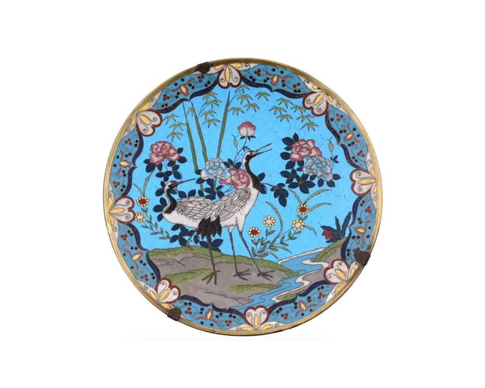 A highly decorative antique Japanese Meiji era brass plate. The exterior of the plate is adorned with a polychrome image depicting two cranes on the bank of a stream surrounded by blooming flowers and bamboo stalks on a turquoise ground made in the