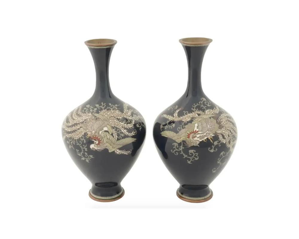 A pair of matching Japanese vases featuring the globular body rising from a short spreading foot to a tall slender neck with flaring rim, decorated in variously coloured cloisonne enamels, depicting a flying phoenix on a deep cobalt blue ground.