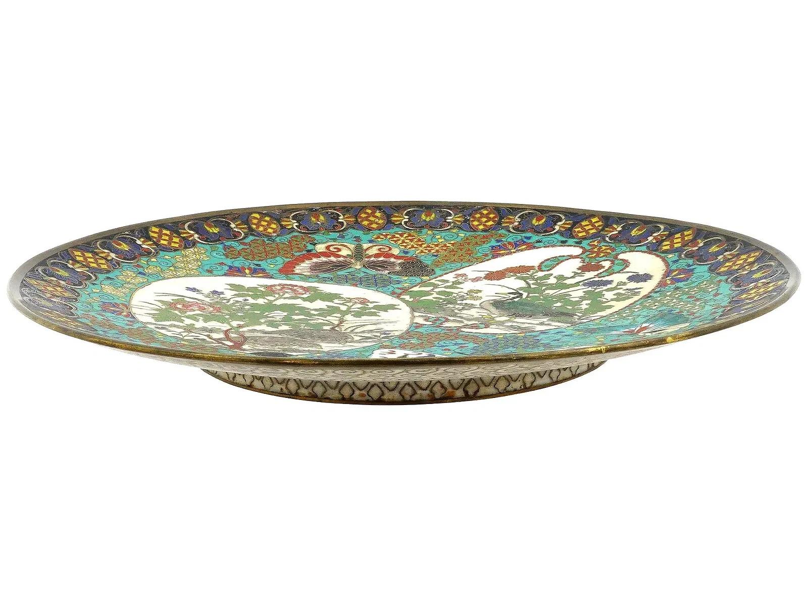 Assiette japonaise ancienne ornée d'un motif en émail cloisonné polychrome. La face avant est décorée d'ornements floraux et de deux médaillons représentant des oiseaux et des branches de fleurs. Les médaillons sont entourés de papillons et de