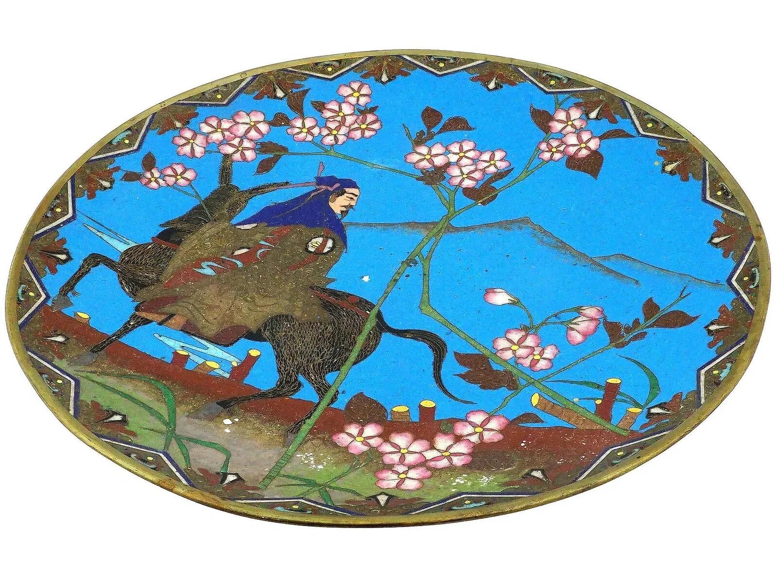 Un antico piatto di rame giapponese del periodo Meiji ornato da un disegno in smalto cloisonne policromo. Ha un colore di fondo blu. Il piatto raffigura un uomo a cavallo, circondato da fiori, su uno sfondo di montagne. Decorato con motivi floreali