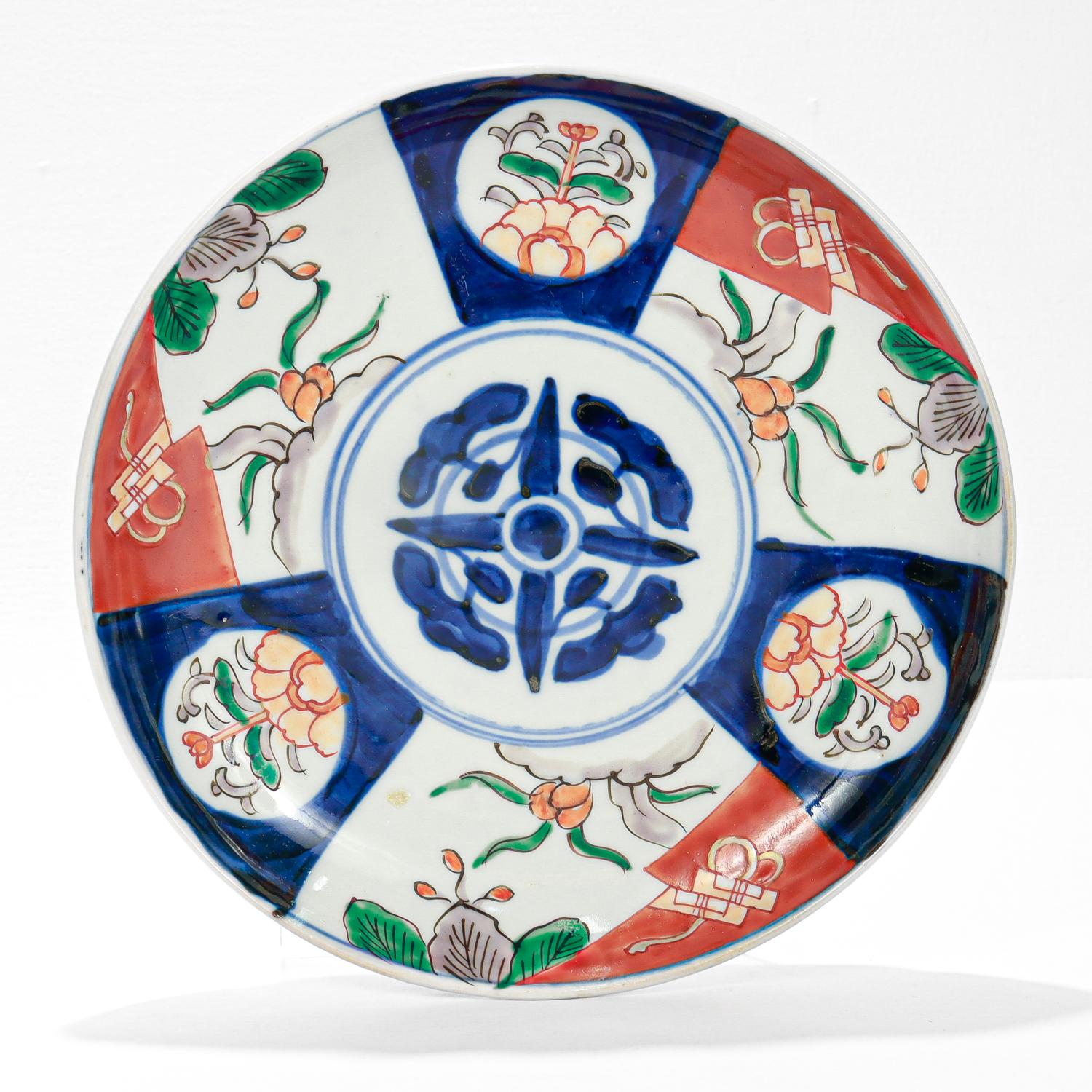 A fine antique Japanese Imari plate.

With a white ground and decorated with geometric and floral devices in blues, reds, oranges, and oranges. The blue cross device to the middle is reminiscent of a shimazu mon.

The exterior of the rim has blue