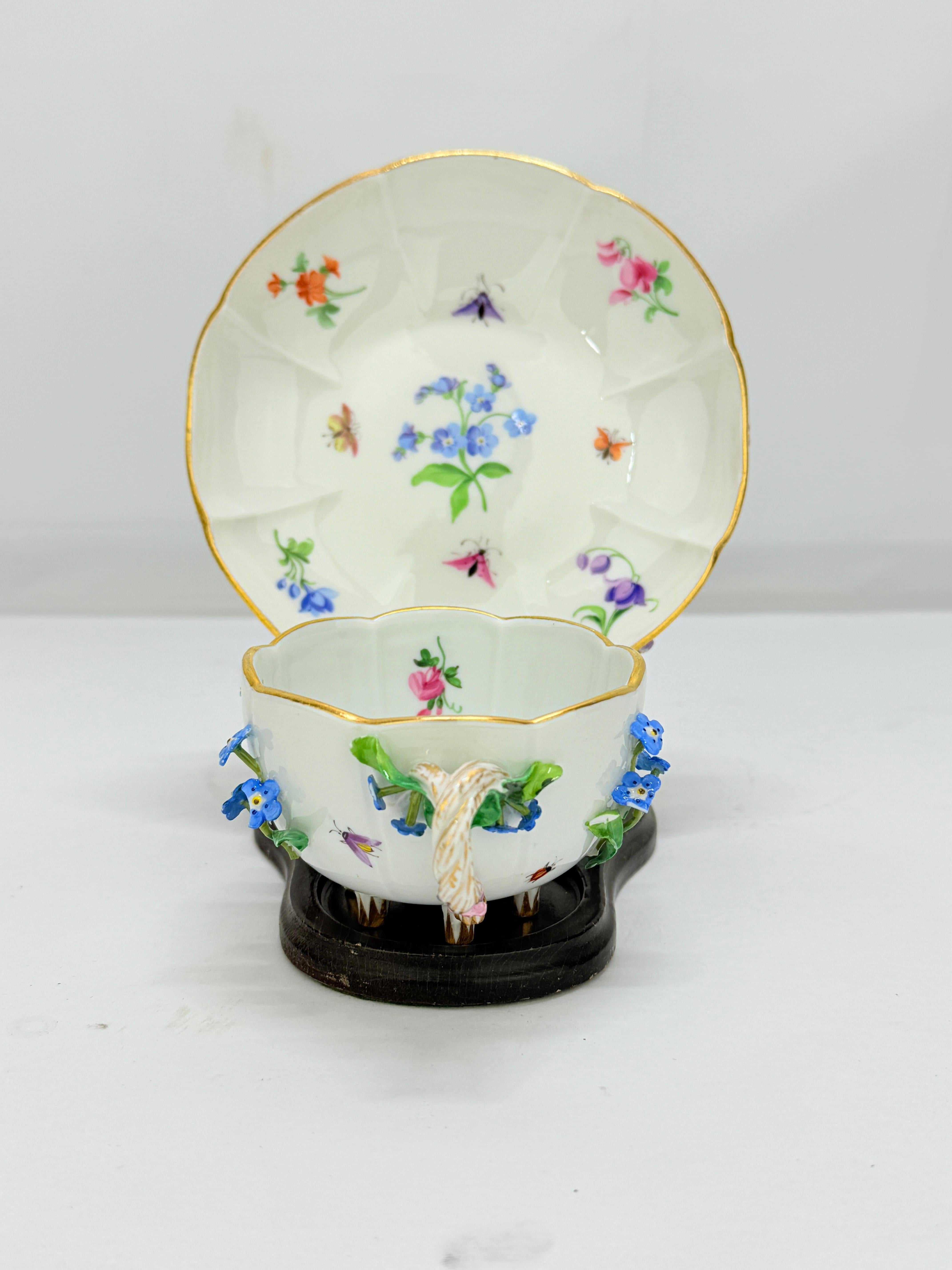   Meissen, Germany, late 19th to early 20th century  
Hard-paste porcelain, hand-painted with polychrome enamels and gilt accents, applied porcelain flowers, five small knop feet. 

This exquisitely detailed porcelain cup and saucer exemplifies the