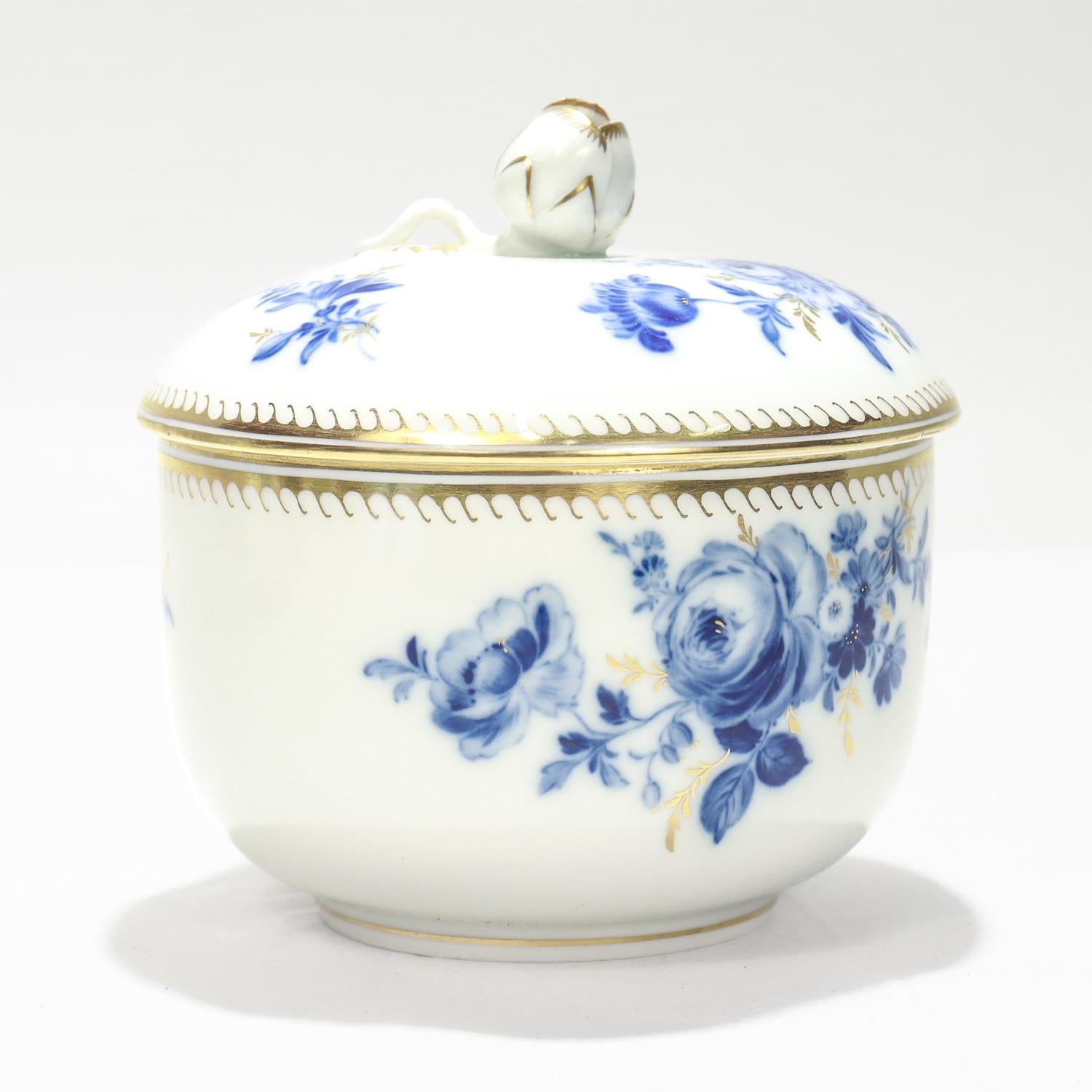 Una fina caja antigua con tapa de porcelana.

Por Meissen.

Decorada con flores azules bajo vidriado con tallos y ramitas dorados sobre fondo blanco, y la tapa con un remate figurado de capullo de flor.

El borde, tanto del cuerpo como de la tapa,