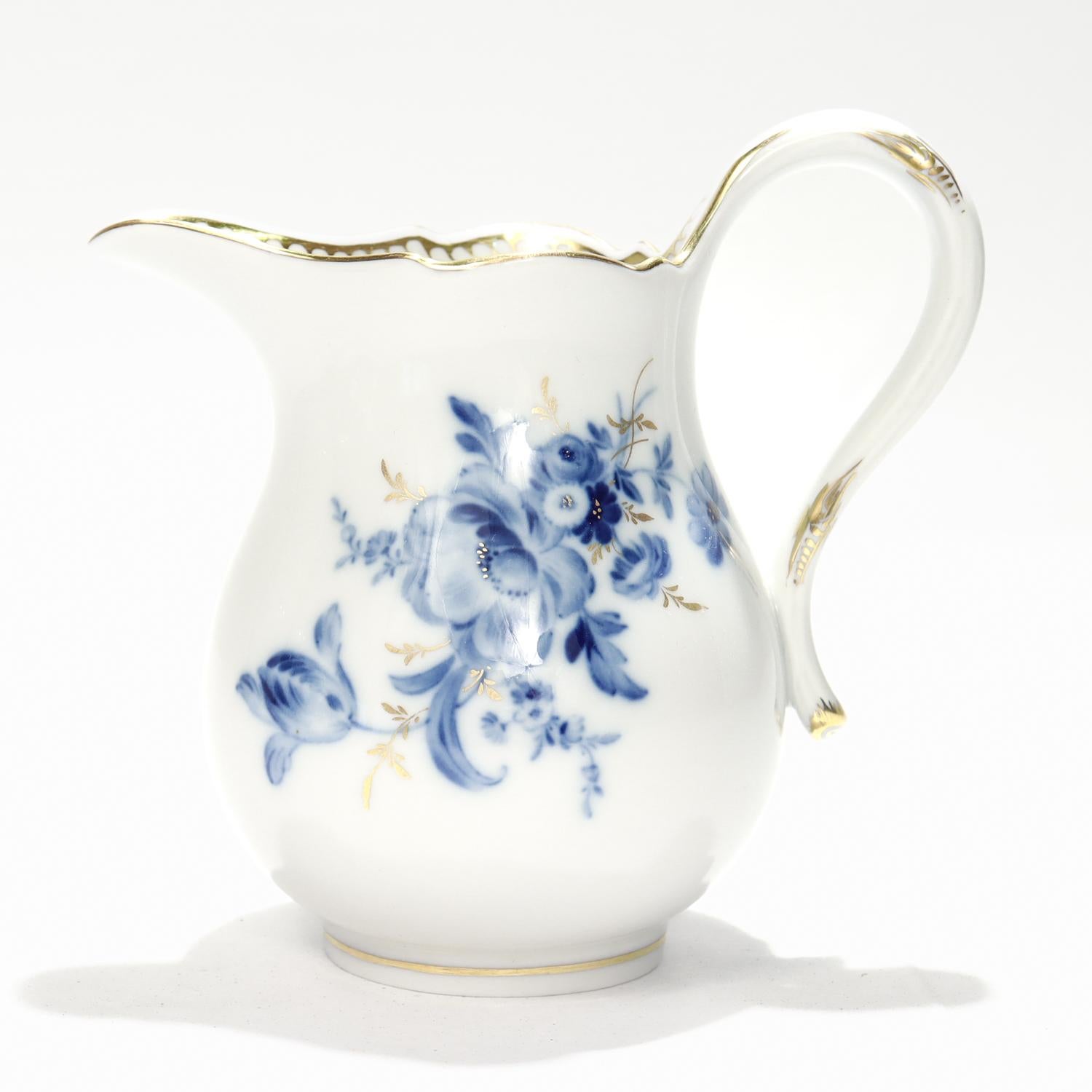 Jarra lechera o cremera de porcelana antigua.

Por Meissen.

Decorado con flores azules bajo vidriado con tallos y ramitas dorados sobre fondo blanco.

El borde interior presenta un dibujo ondulado dorado en la pared interior. El asa y el borde del