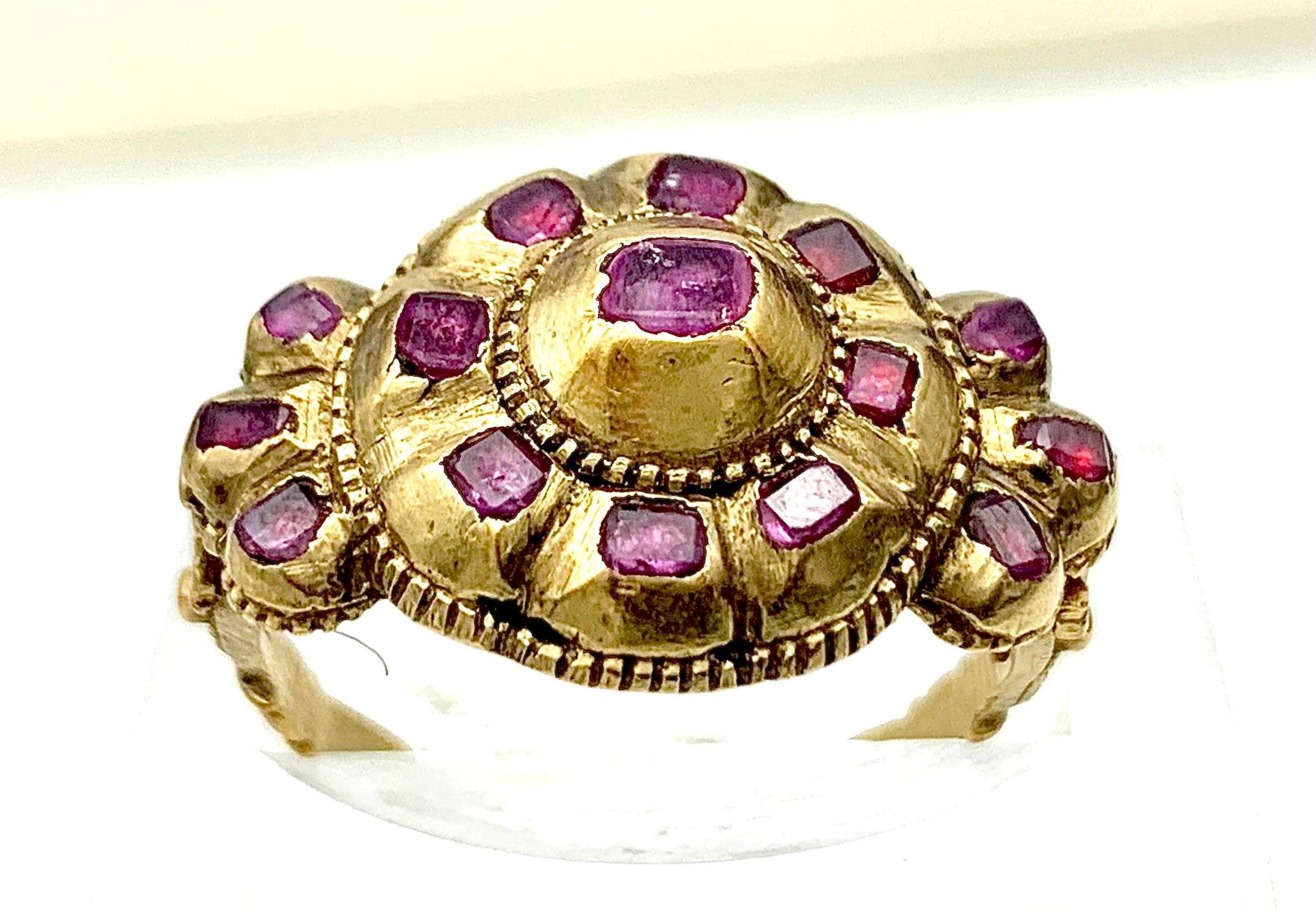 This mid-eighteenth century ring is set with flat cut rubies. The ring head in the shape of a rosette is set with eight small and one large ruby. The larger center stone in a dome shaped closed mount is riveted to the ring head.  each ring shoulder