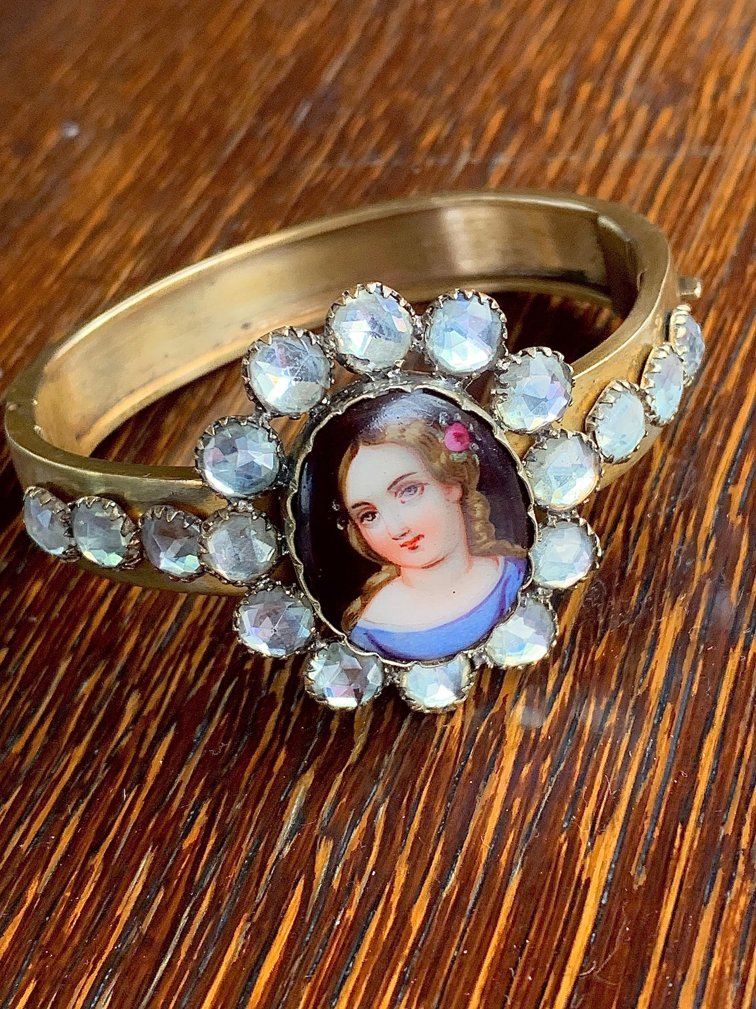 Questo affascinante braccialetto è stato creato nel 1850 ca in metallo dorato. Il pezzo centrale è una targa ovale convessa in porcellana con l'immagine stampata di una ragazza con un vestito blu e una rosa tra i lunghi capelli biondi e ricci. La