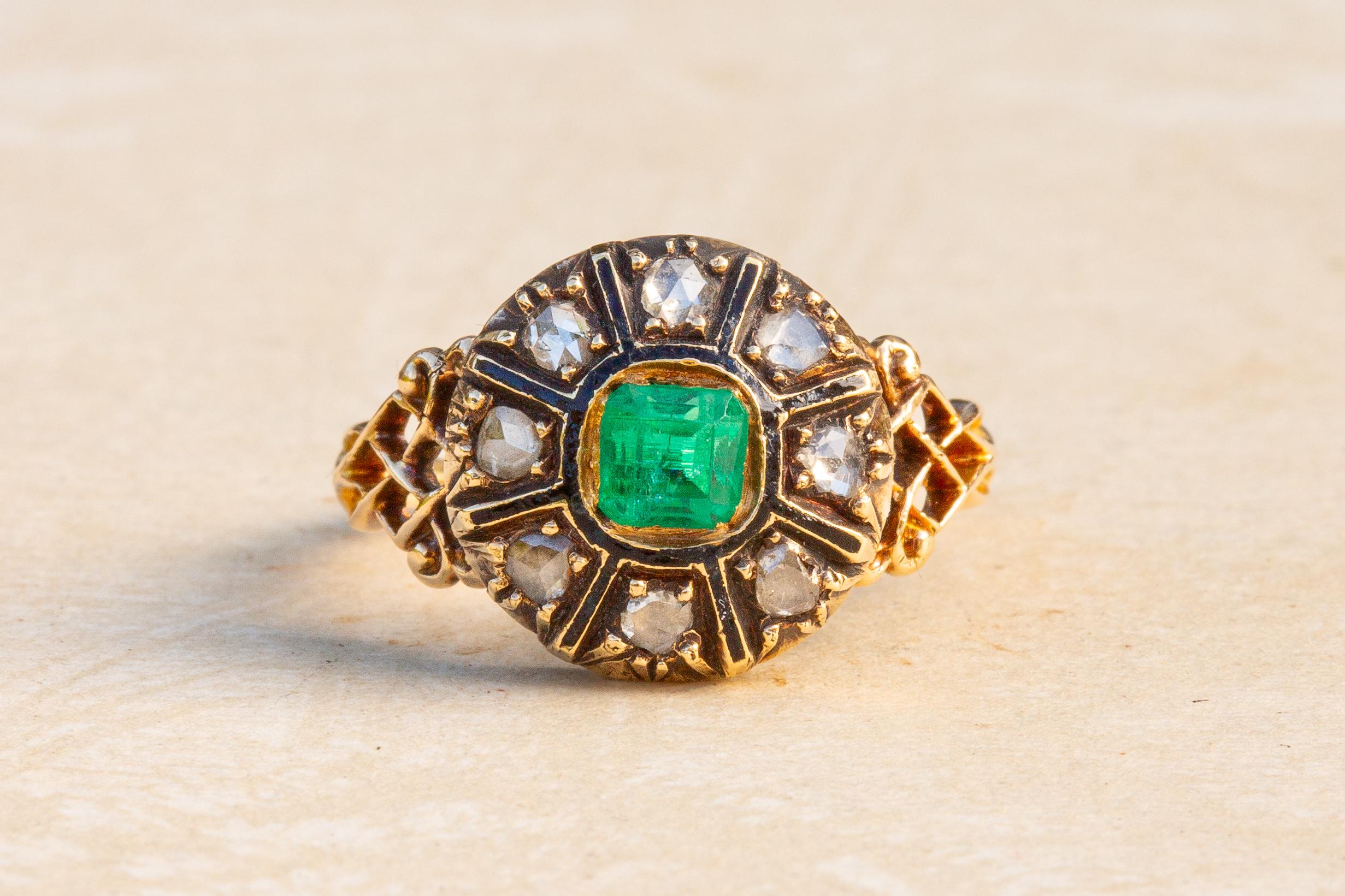 This striking antique gold cluster ring dates to the mid-19th century. 

The ring is crafted in 18K gold with black enamel. In the centre rests a vibrant 0.44ct step cut emerald in an open-backed rubover setting. The stone is a wonderful vivid green