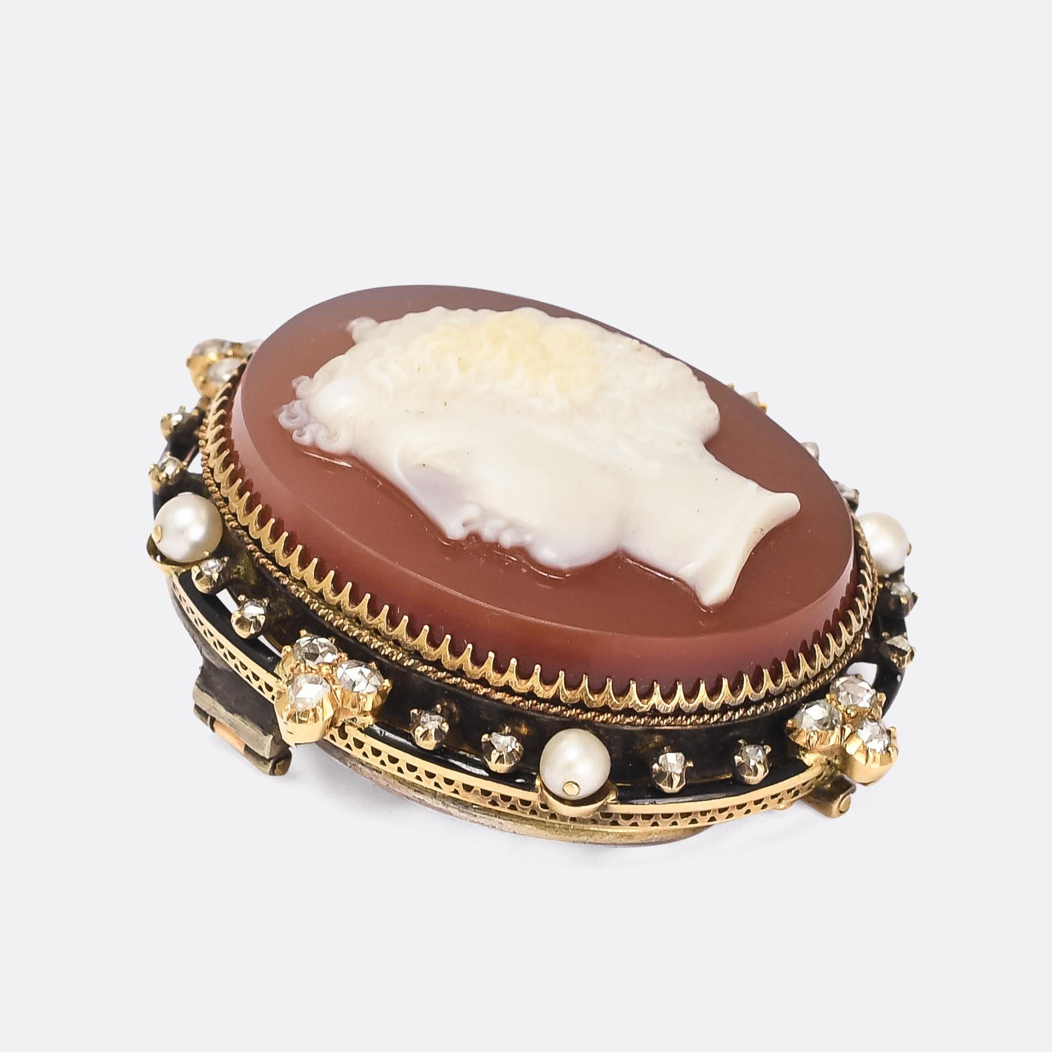 A cool Victorian cameo brooch mounted in an elaborate rose cut diamond, natural pearl, and black enamel border. The cameo itself is wonderfully executed, in white hardstone agate with a red backdrop, it depicts a classical style woman with tightly