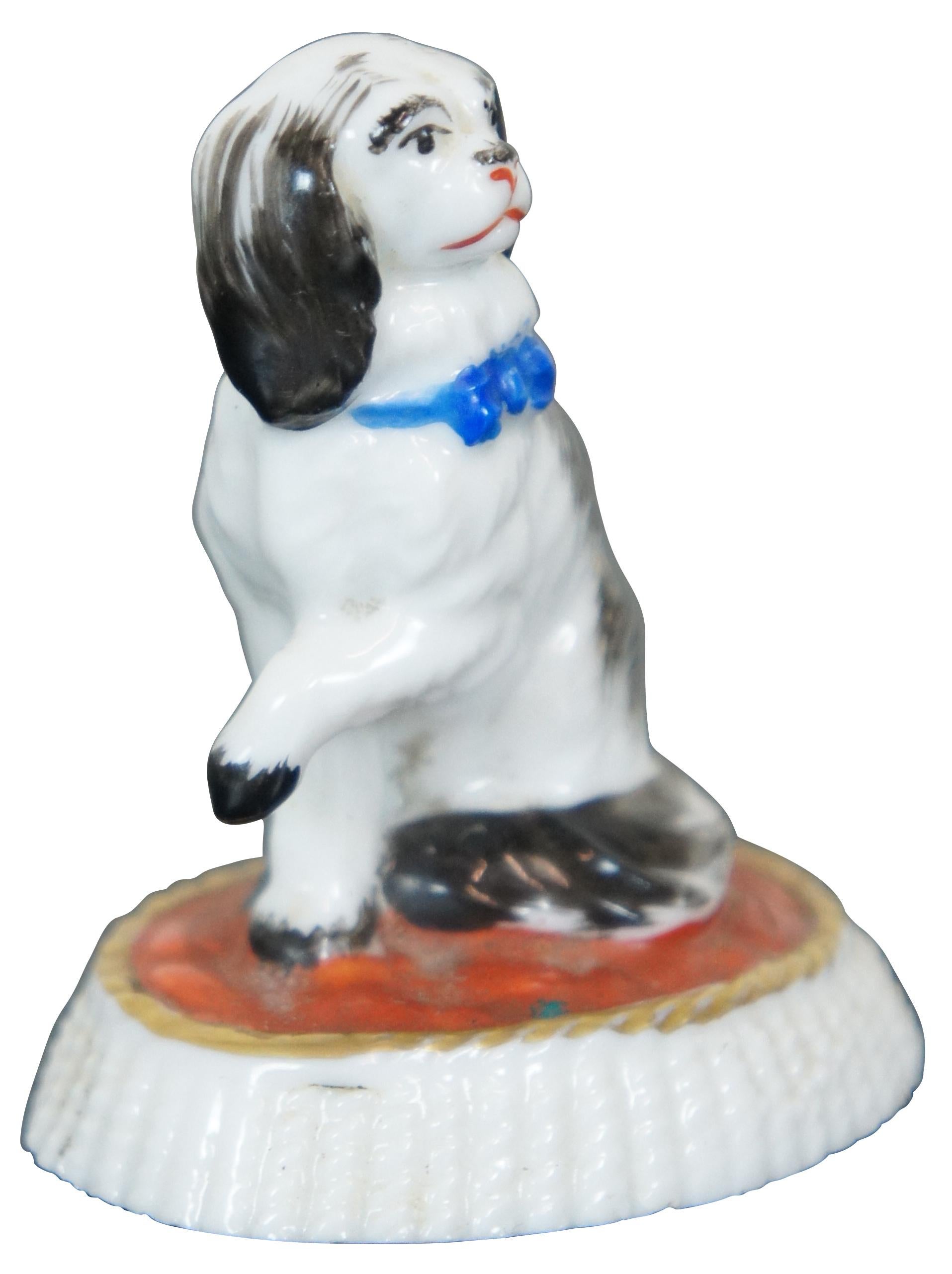 Antique 1885s - 1920s century miniature porcelain figurine in the shape of a black and white spaniel with a blue bow, seated on a red and white fringed cushion. Marked with a Samson golden anchor. This mark was used on German Chelsea Reproductions